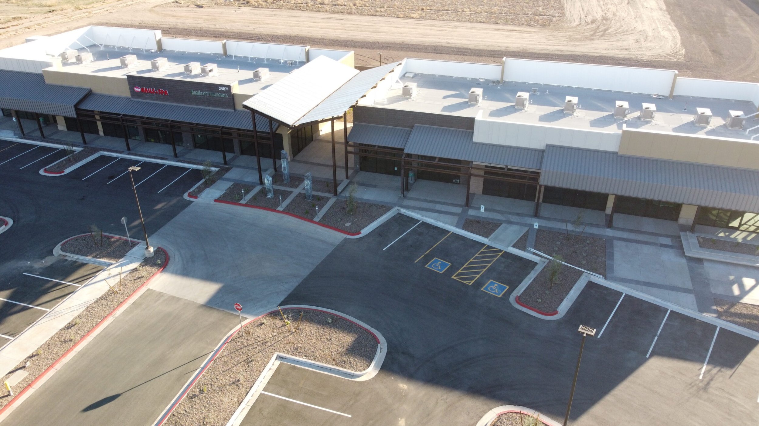 An aerial view of a mostly empty commercial building parking lot with designated wheelchair accessible parking spaces, lamp posts, and some small landscaped areas with a few young trees.