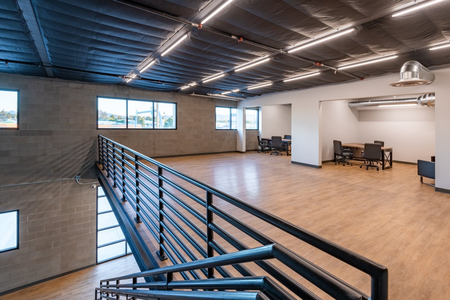 Empty modern office space with wooden floors, large windows, black metal railing, white walls, and nearby work desks with chairs.