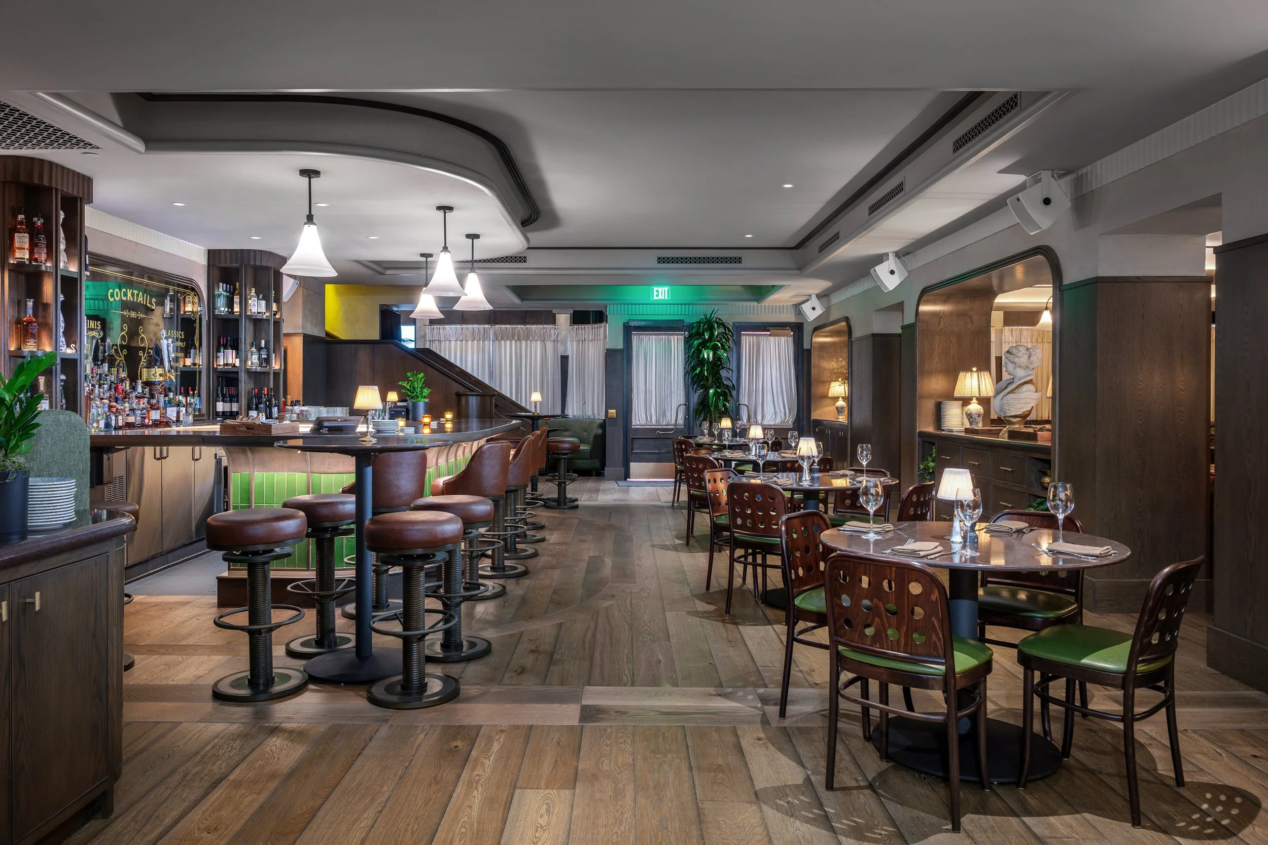 Interior of a restaurant or bar with wooden furniture, tables set with glasses and napkins, bar area with stools, and ambient lighting.