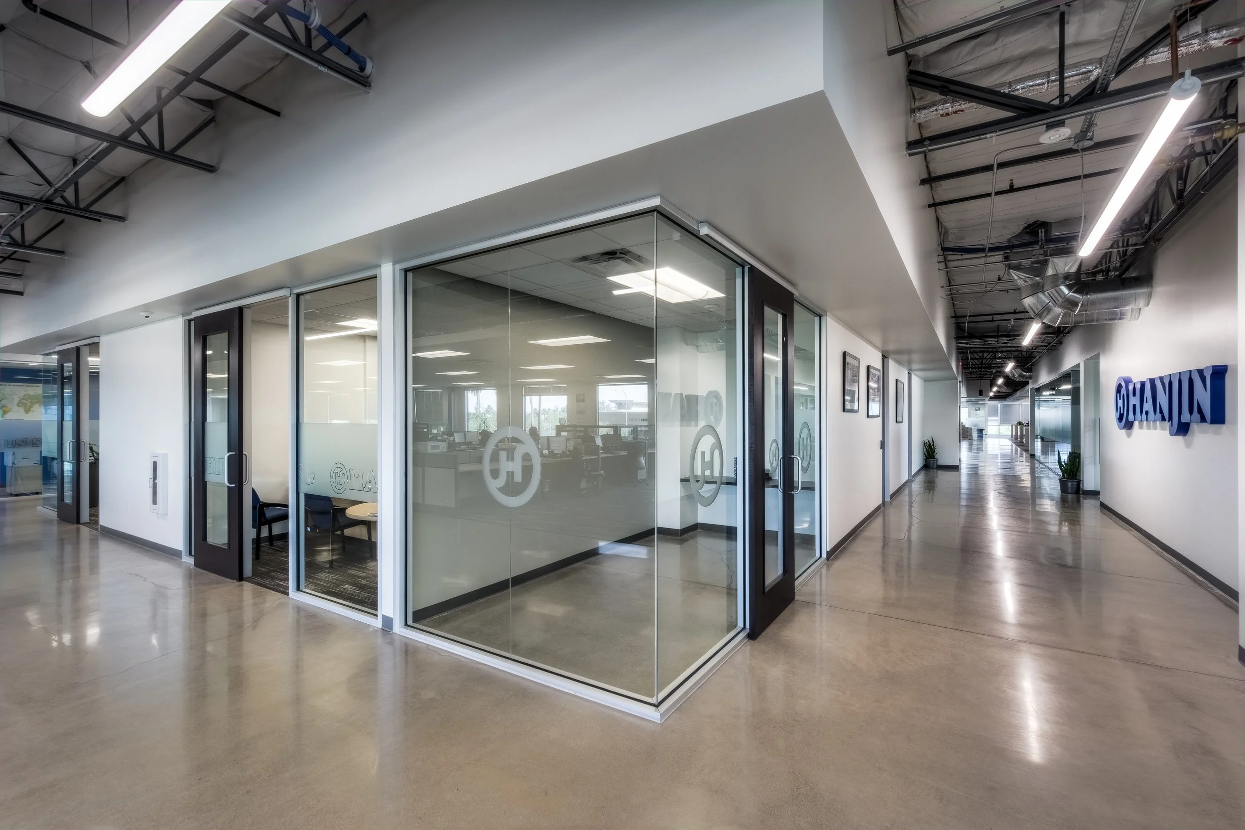 Modern office hallway with glass enclosed conference room, for CHAININ, on the right side of the hallway. The hallway has polished concrete floors, plants, framed artwork, ceiling with exposed ductwork, and contemporary lighting.