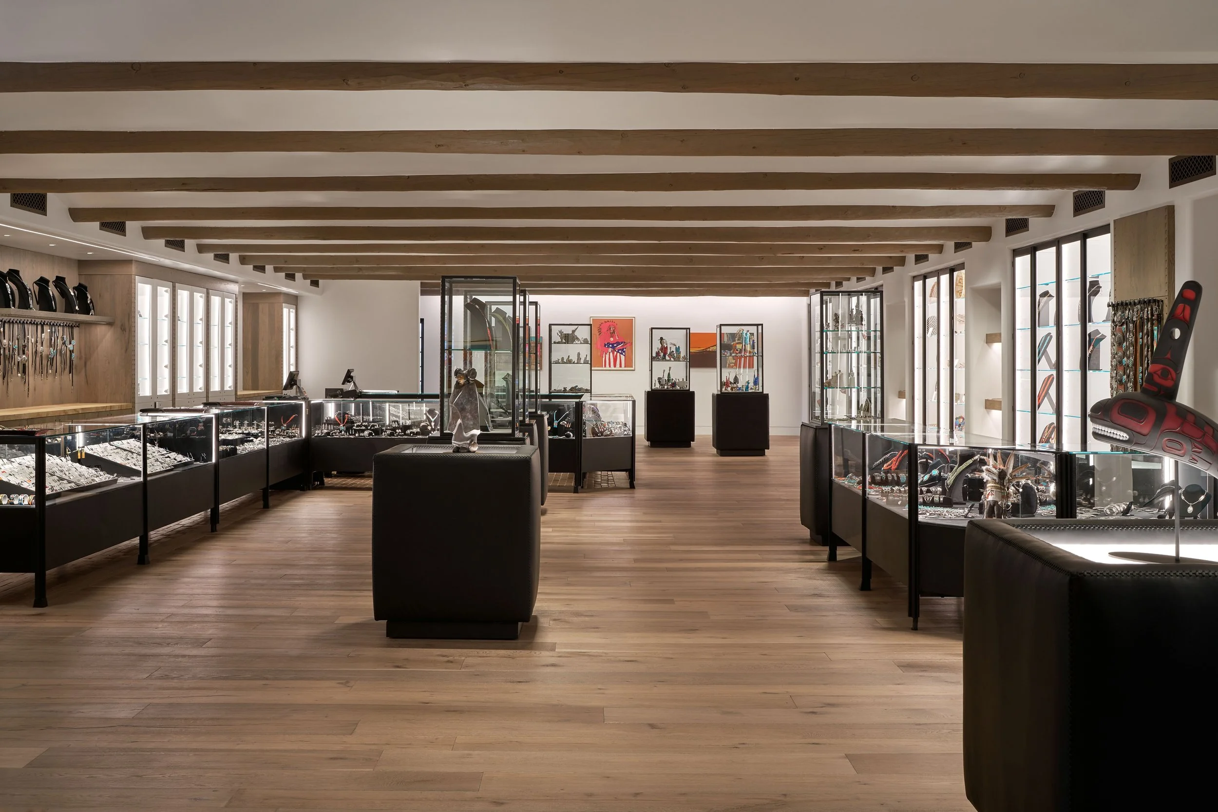 Interior of a jewelry store with display cases filled with jewelry and accessories, wooden floors, and ceiling beams, with light coming through large windows.