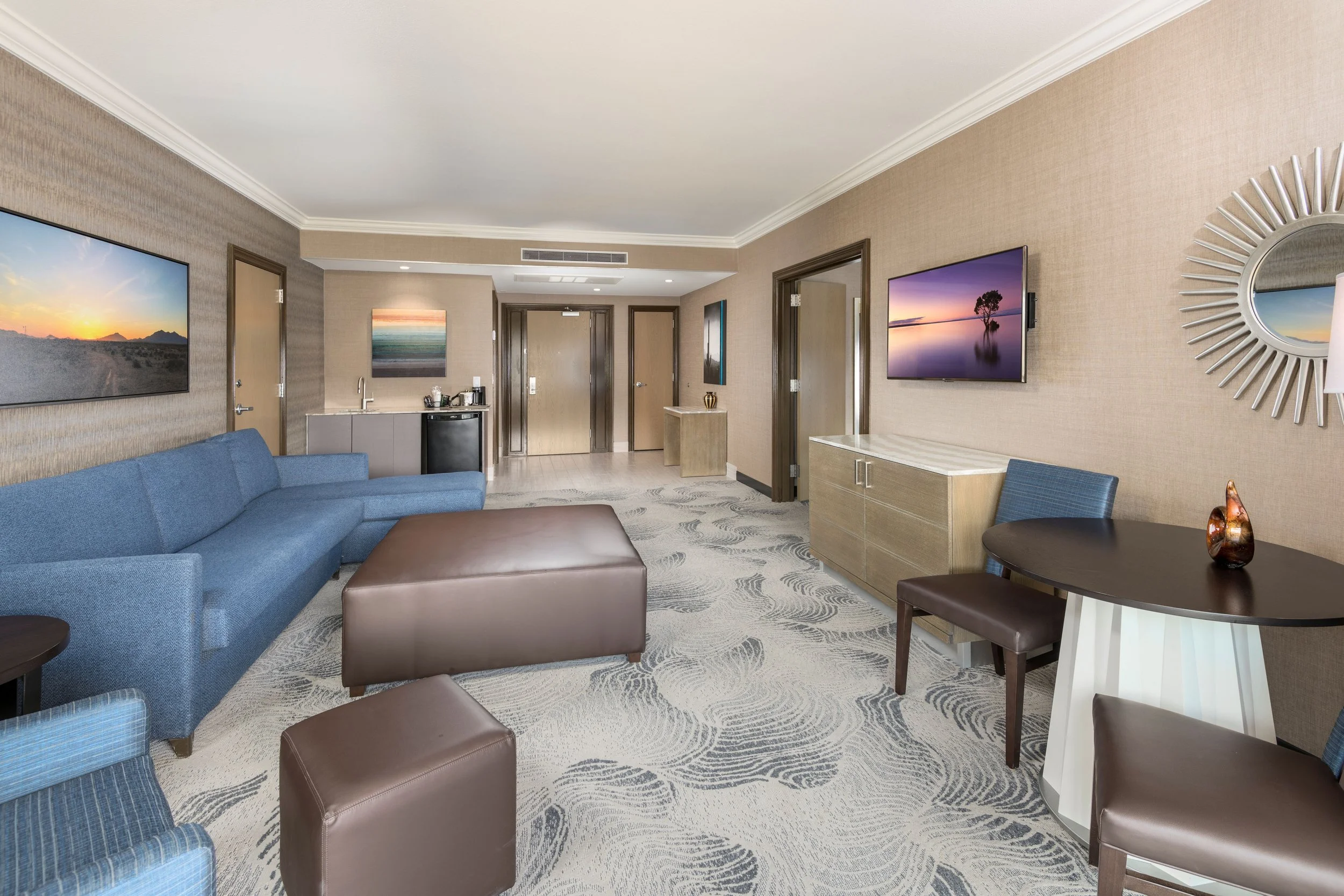 Hotel suite living room with a blue couch, brown ottomans, wall-mounted TVs, artwork, and seating area.