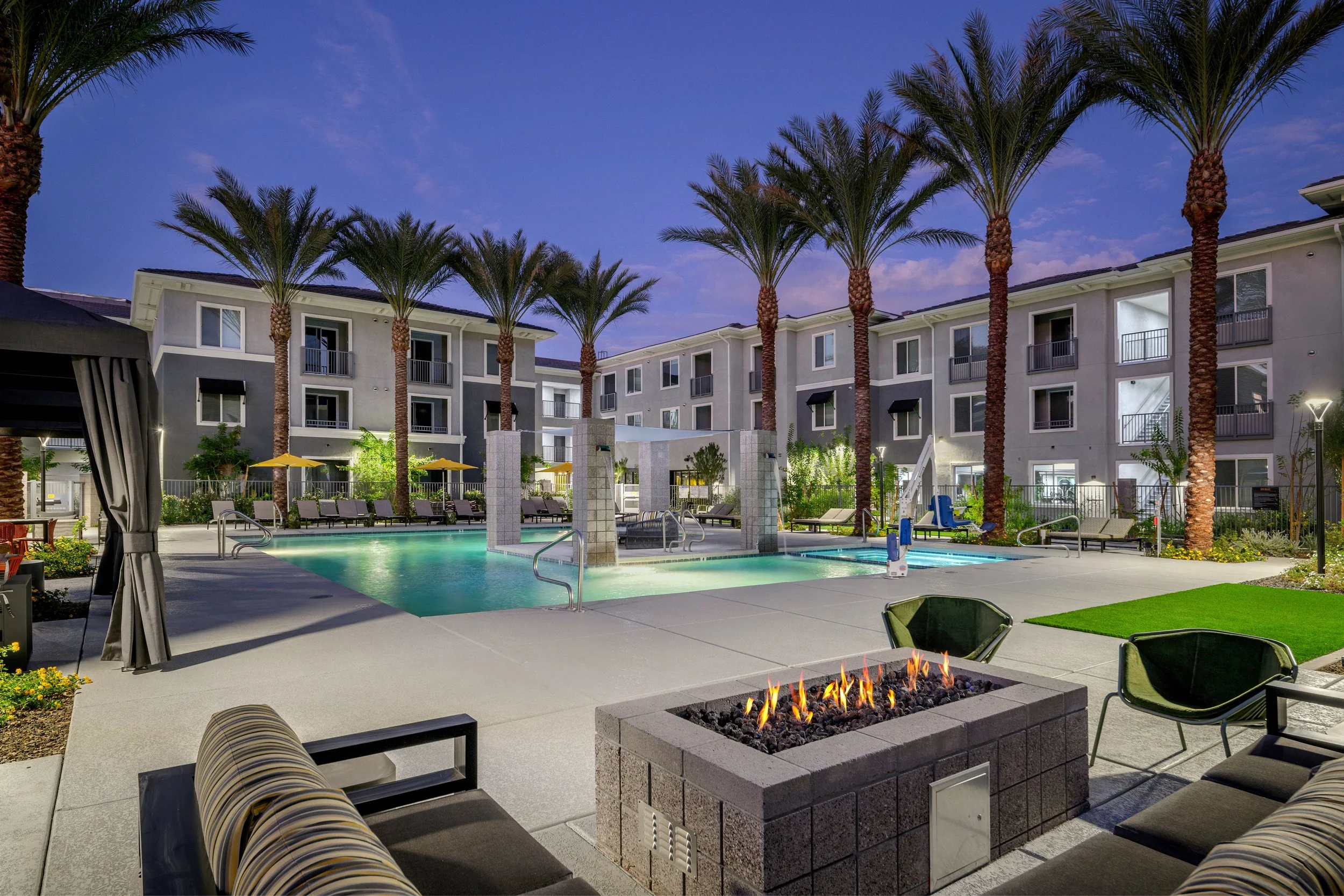 Night view of a modern apartment complex with a swimming pool surrounded by lounge chairs, palm trees, and a fire pit in the foreground.