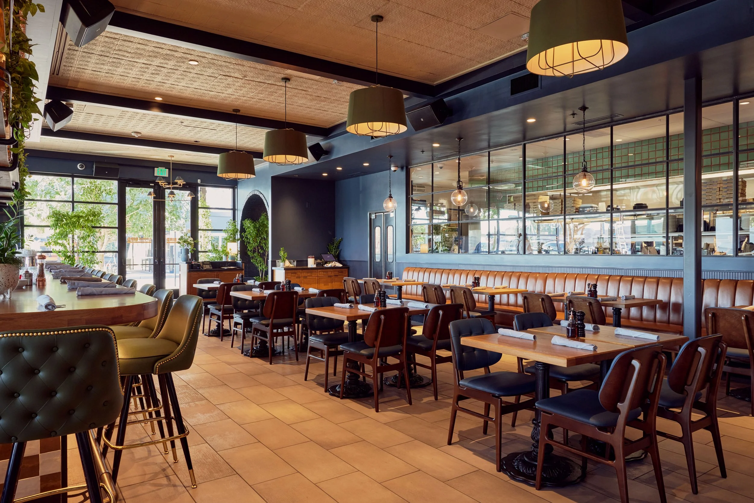 Interior of a modern restaurant with wooden tables, black and brown chairs, and a leather bench along the wall. Green pendant lights hang from the ceiling, and large windows let in natural light. Some potted plants are near the entrance.