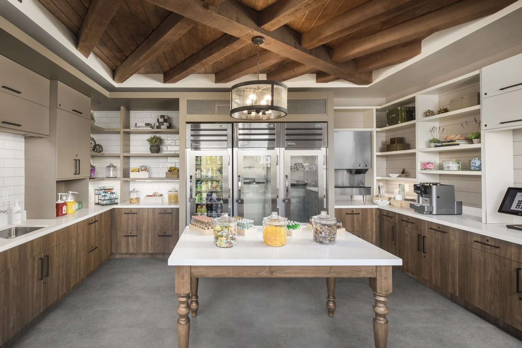 A modern kitchen with wooden cabinetry and a white countertop island in the center. Open shelves and a freezer behind glass cabinets are visible. The ceiling features wooden beams and a hanging circular light fixture.