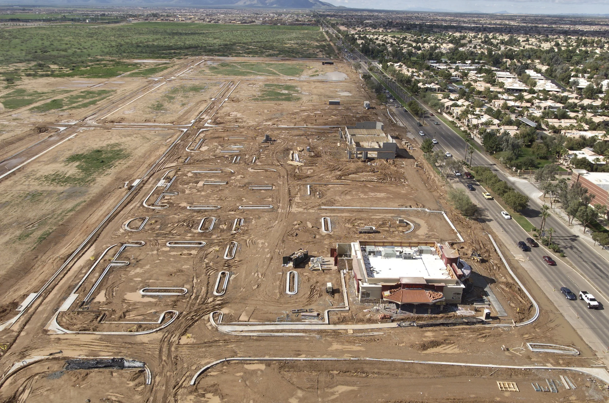 An aerial view of a construction site with foundational pipelines and a partially built building, next to a busy road with cars, residential area, and distant mountains.
