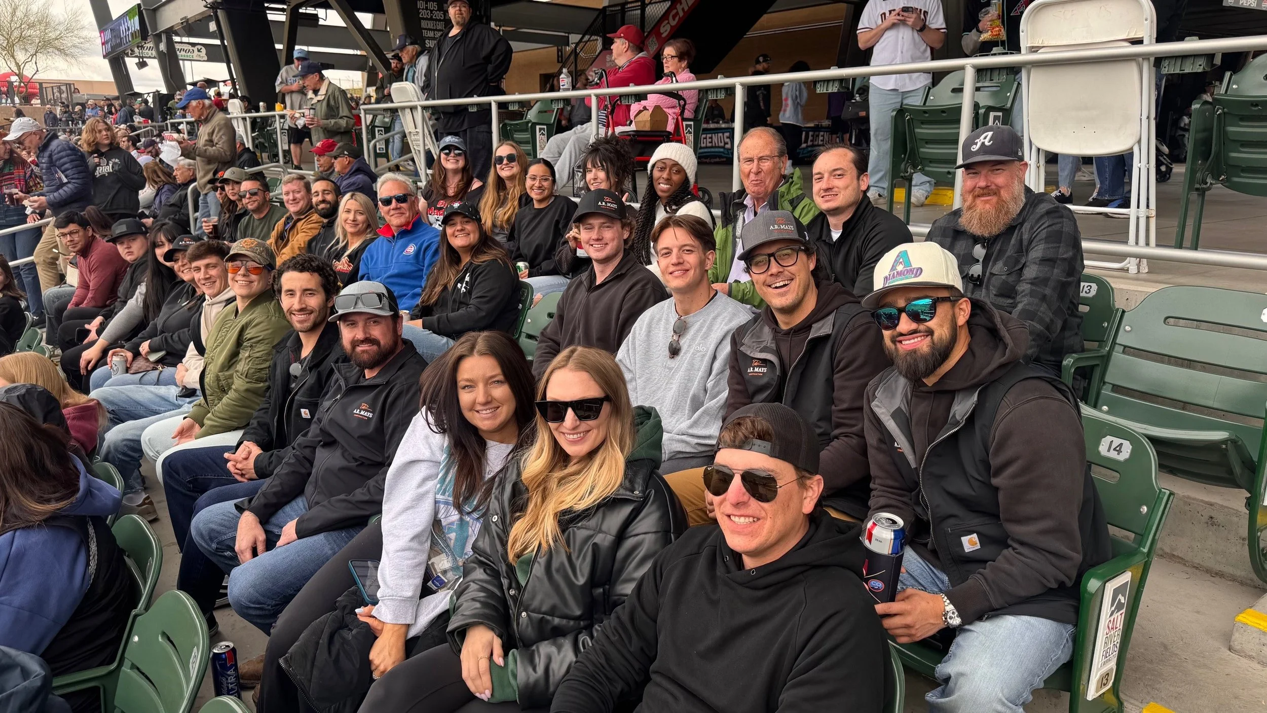 People seated in stadium seats attending an outdoor event, with a diverse group smiling and enjoying the occasion.
