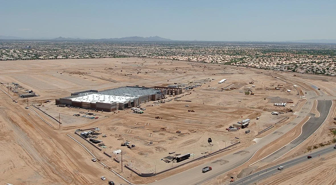 Aerial view of an under-construction commercial area in a desert landscape, with a large building partly completed, surrounding roads, construction vehicles, and a distant city skyline.