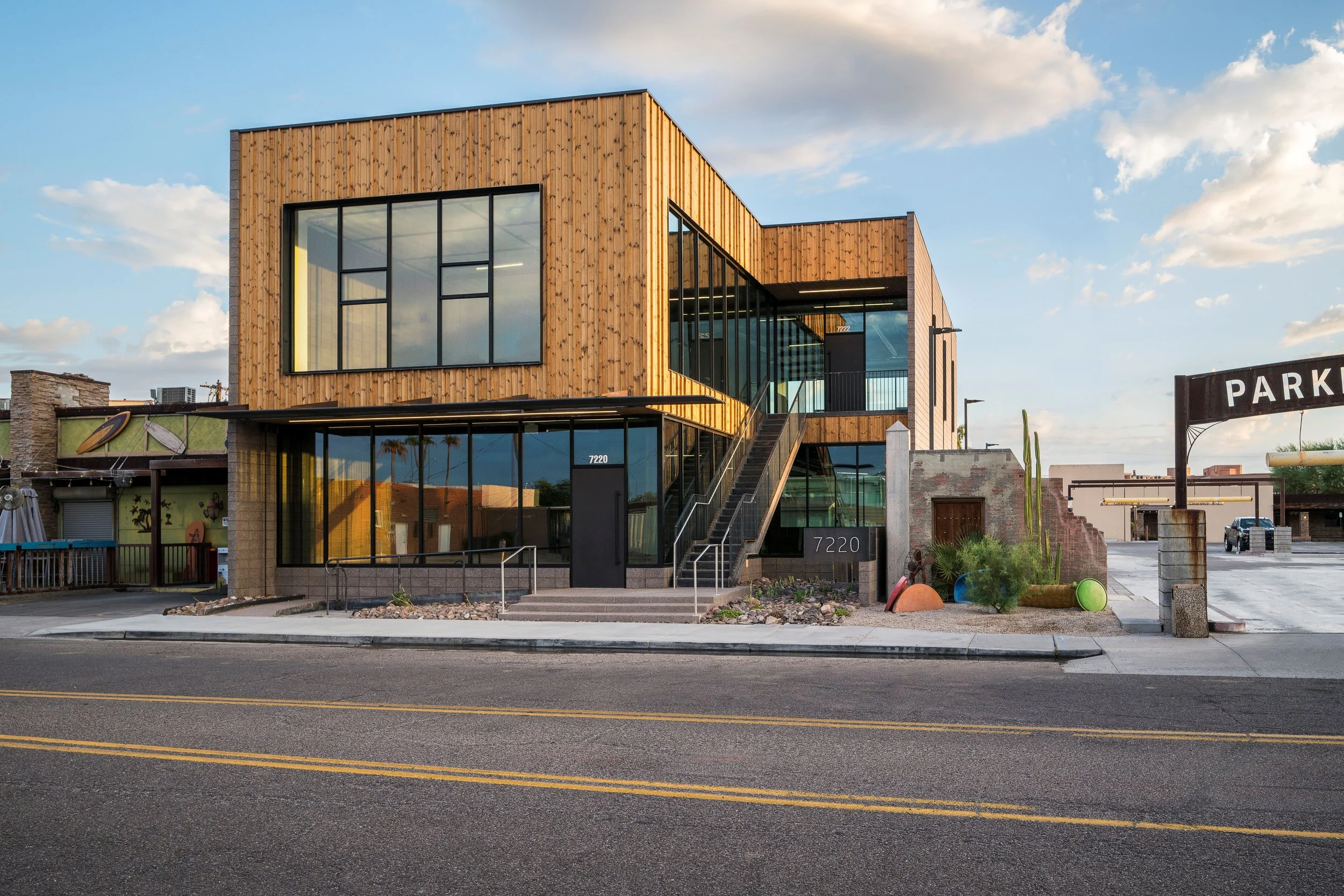 Modern two-story commercial building with wooden and glass exterior, large windows, stairs leading to the entrance, and a parking lot area with desert plants and cactus decor.