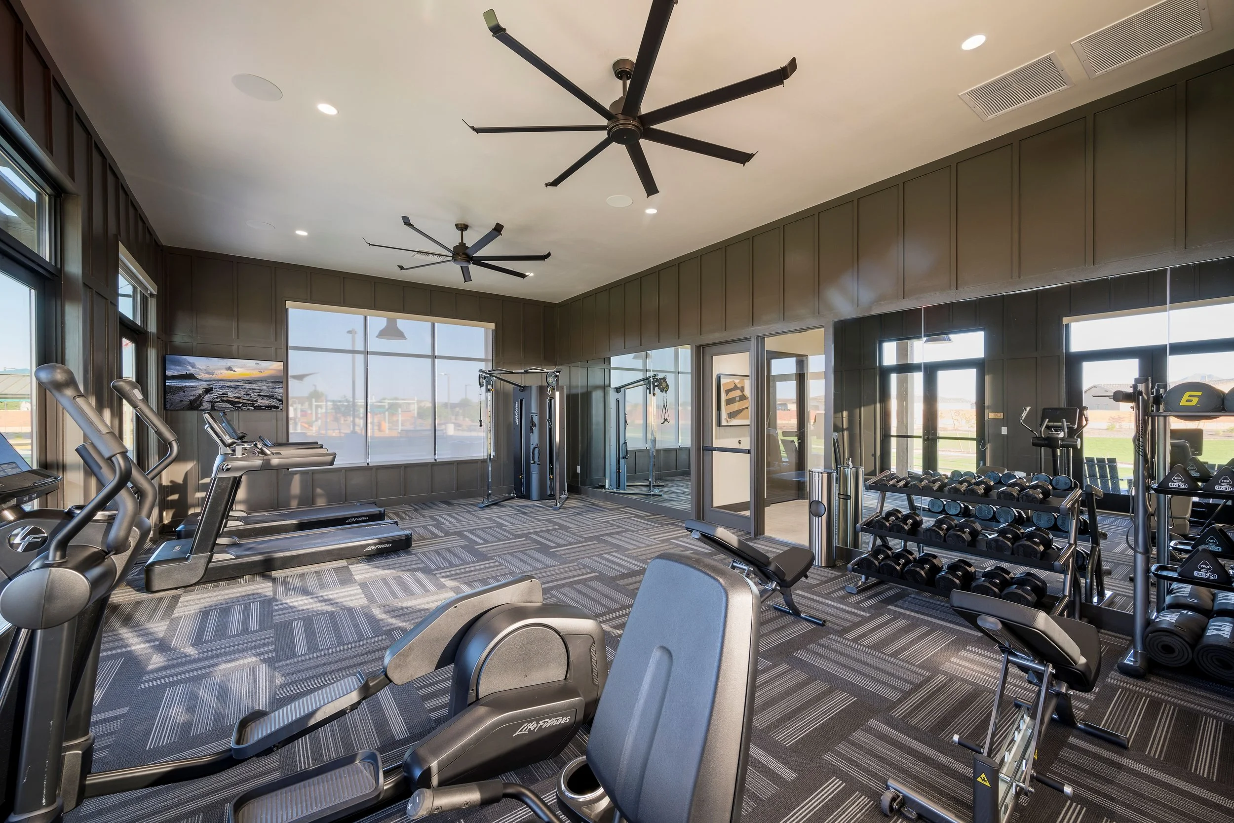Gym with cardio machines, free weights, and exercise equipment, large windows, ceiling fans, and a television.
