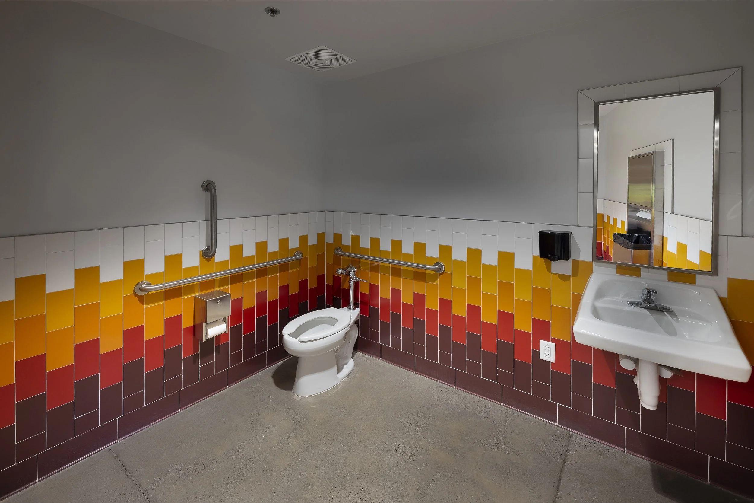 Accessible bathroom with colorful tile design, a toilet with grab bars, a wall-mounted sink, a mirror, and a paper towel dispenser.