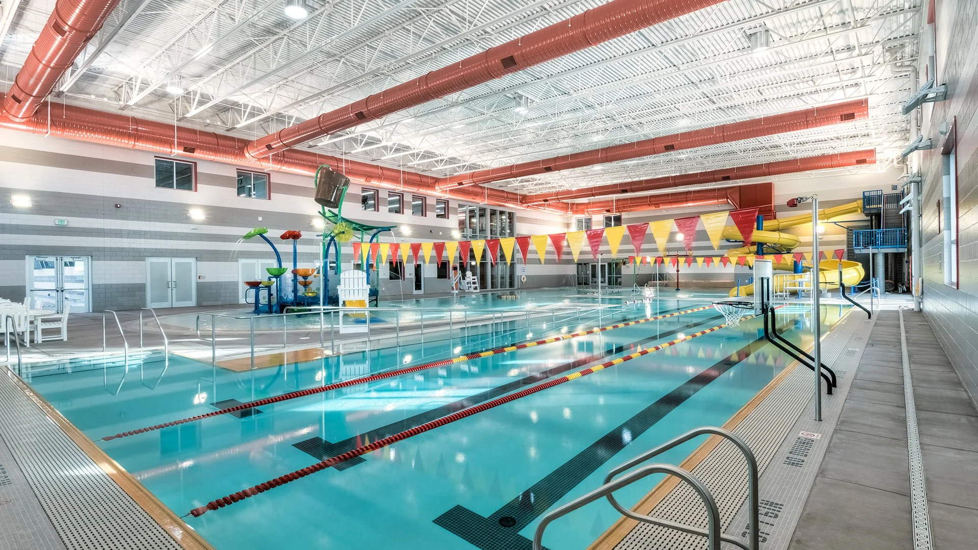 Indoor swimming pool with water slides, pool lanes, and a water play area with water jets, inside a modern aquatic center with bright lighting and large windows.