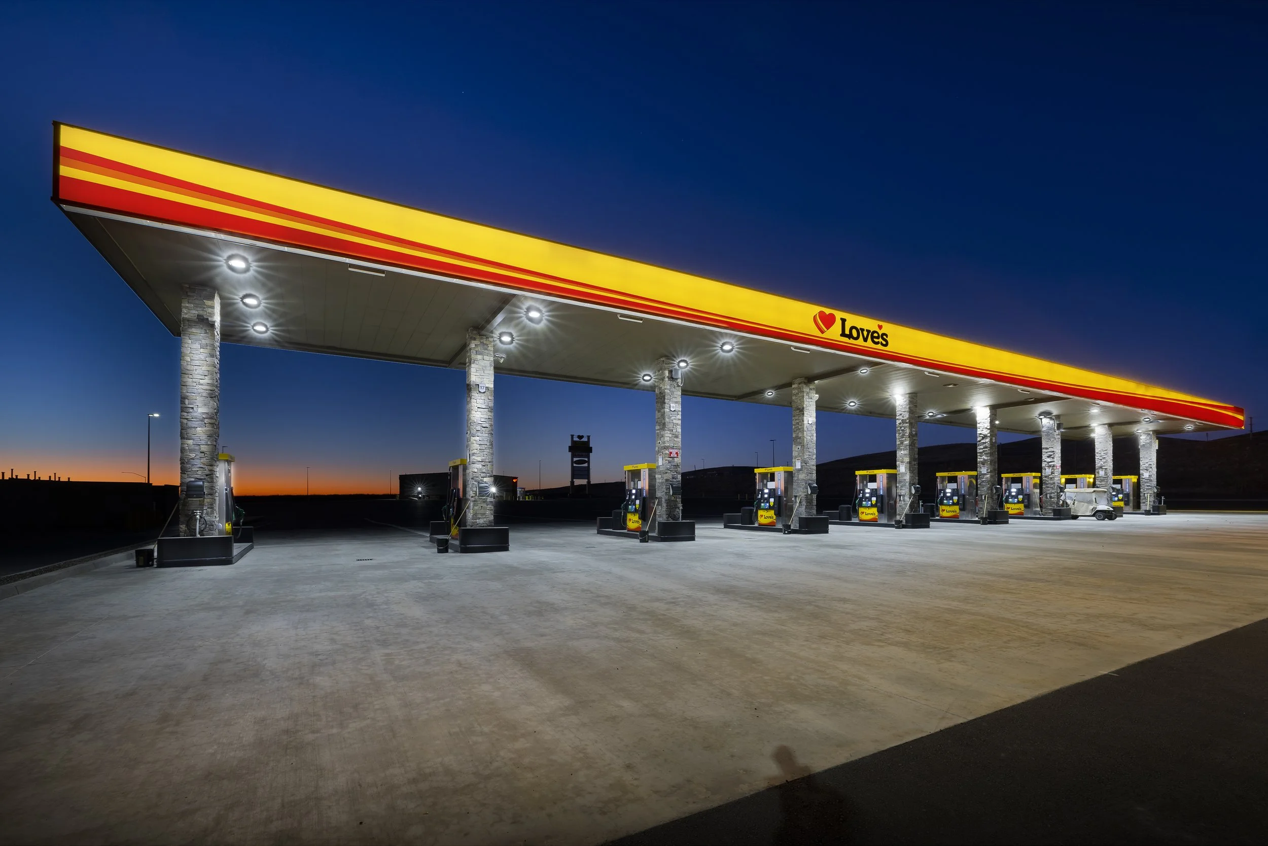 Nighttime view of a Love's gas station with multiple fuel pumps under a large yellow canopy, illuminated by bright lights, with a dark sky and orange sunset in the background.