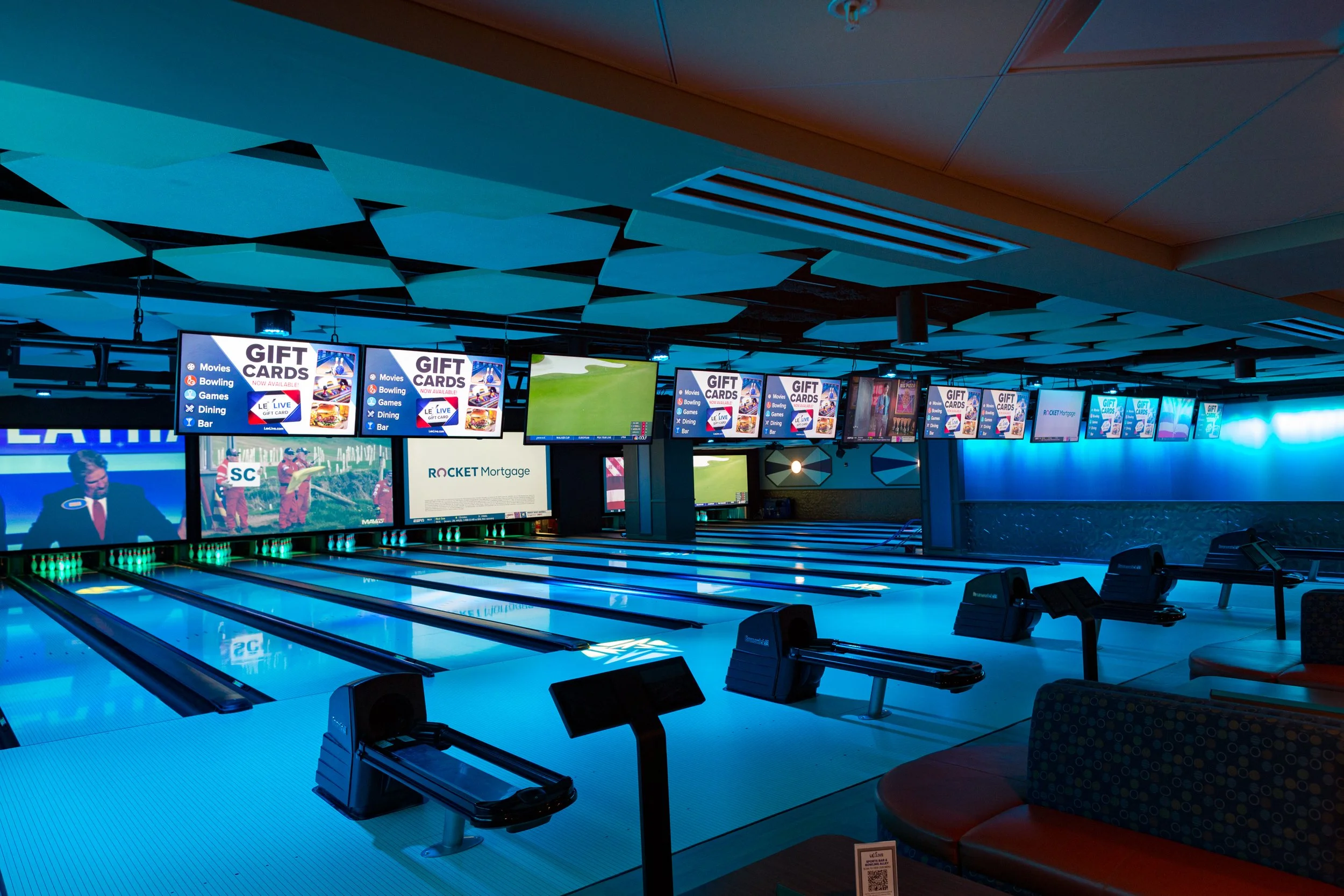 Interior of a modern bowling alley with several lanes, large screens displaying advertisements and game scores, and colorful lighting.