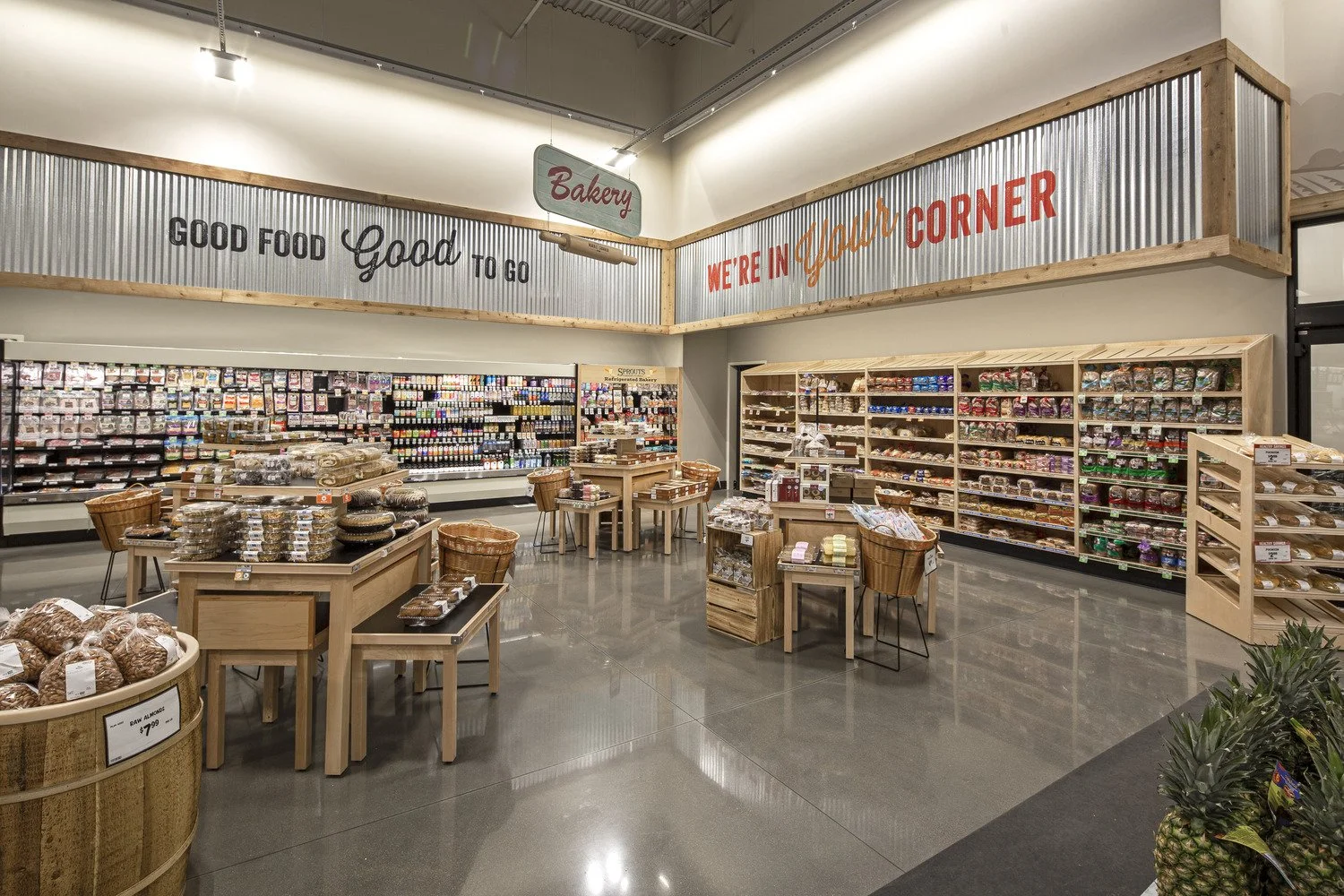 Interior of a bakery section in a grocery store with wooden tables and baskets displaying baked goods, shelves stocked with packaged food items, and signs that read 'Good Food Good To Go' and 'We're In Your Corner' in large letters.
