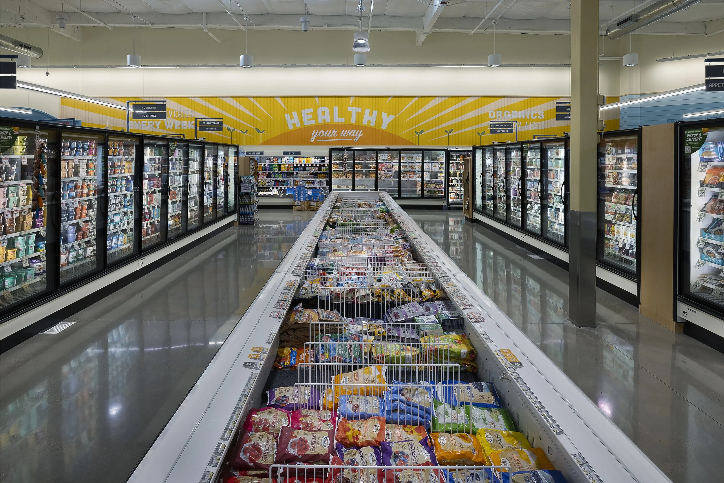 View of a grocery store aisle with a frozen food section on the left and right sides, and an open freezer with various frozen products in the center. A yellow sign in the background reads "Healthy your way".