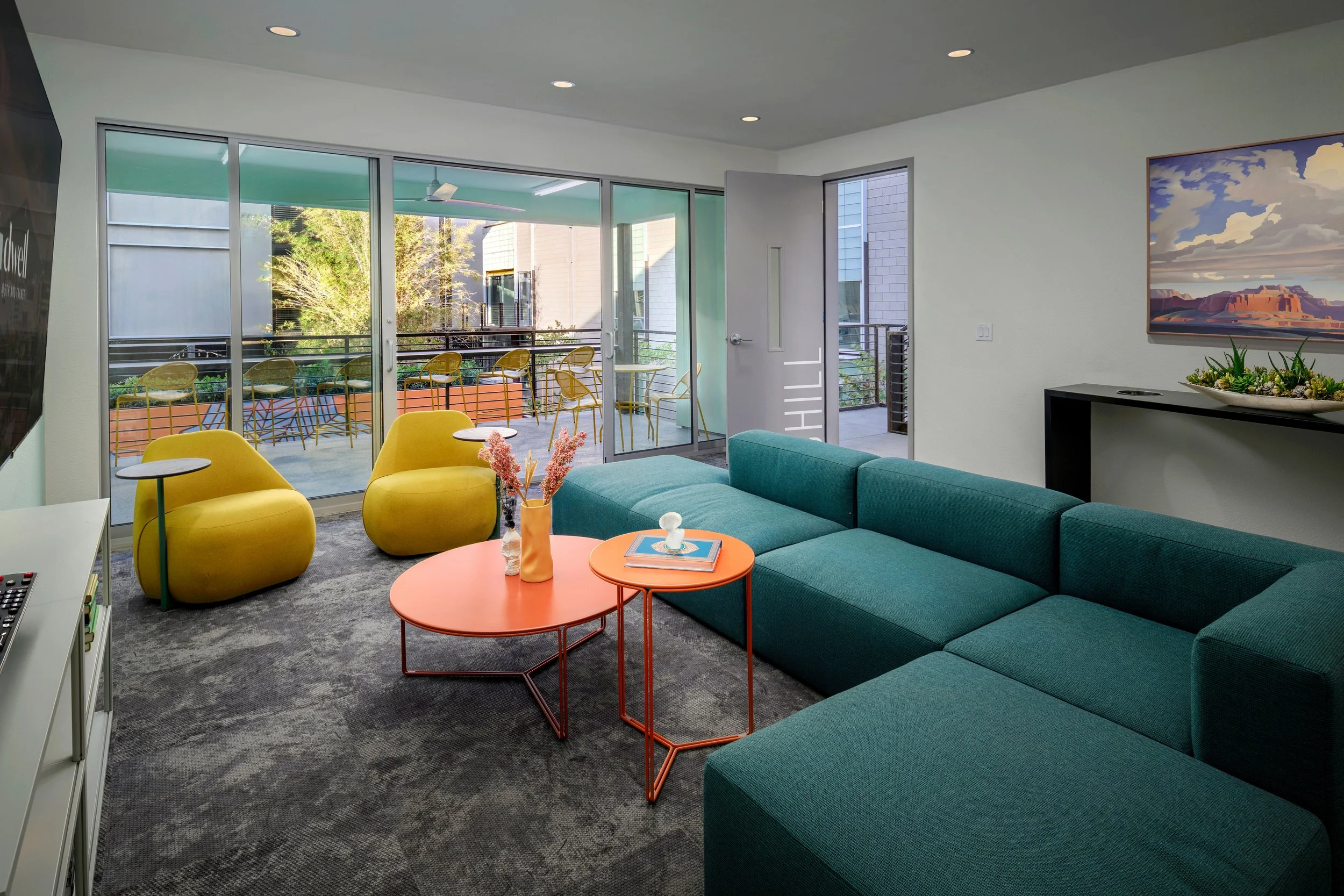 Modern living room with teal sectional sofa, yellow lounge chairs, orange coffee tables with vases and books, sliding glass door leading to balcony with bar stools, wall-mounted television, and mountain landscape picture on the wall.