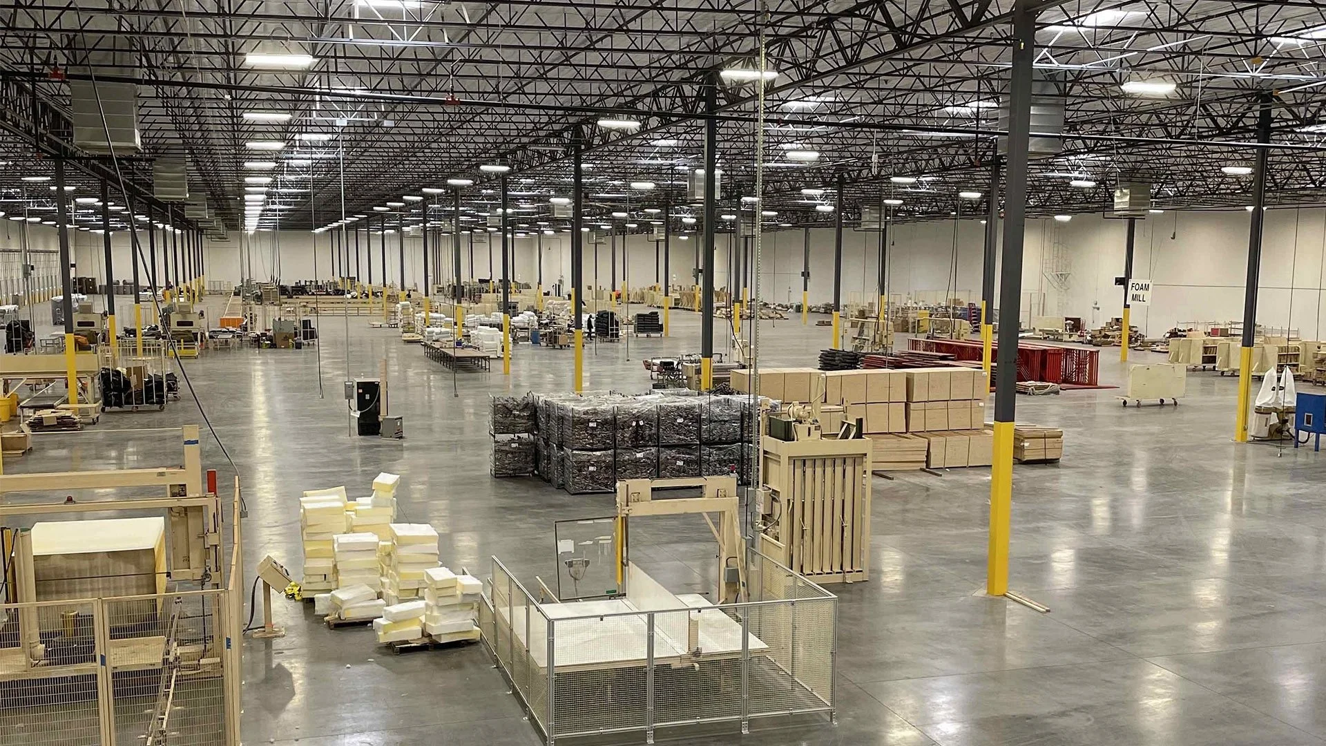 Large warehouse with high ceilings, concrete floor, and scattered pallets of packed boxes, foam, and equipment.