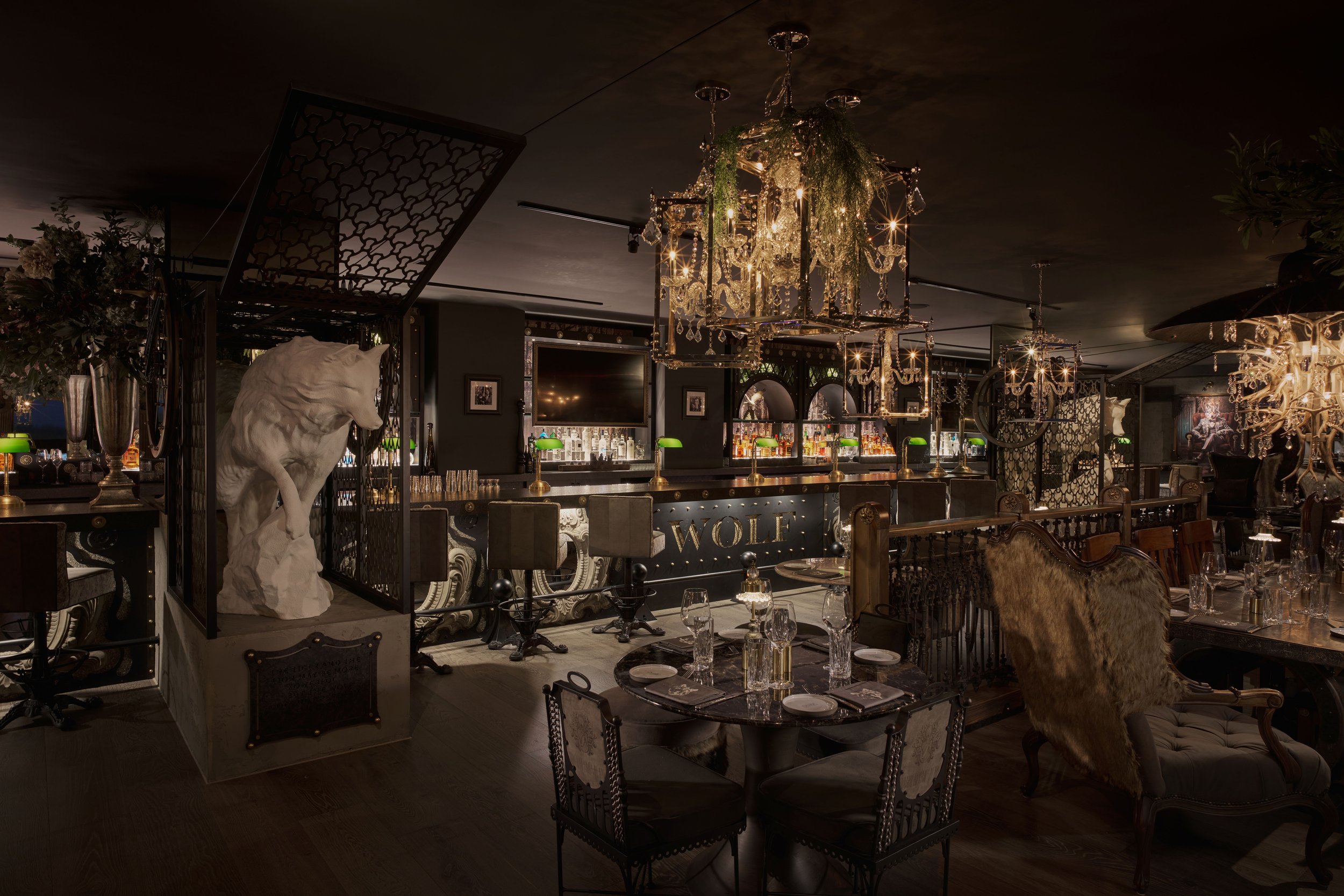 Elegant dimly lit bar and restaurant with crystal chandeliers, antique-style furniture, and a sculpture of a wolf on display.