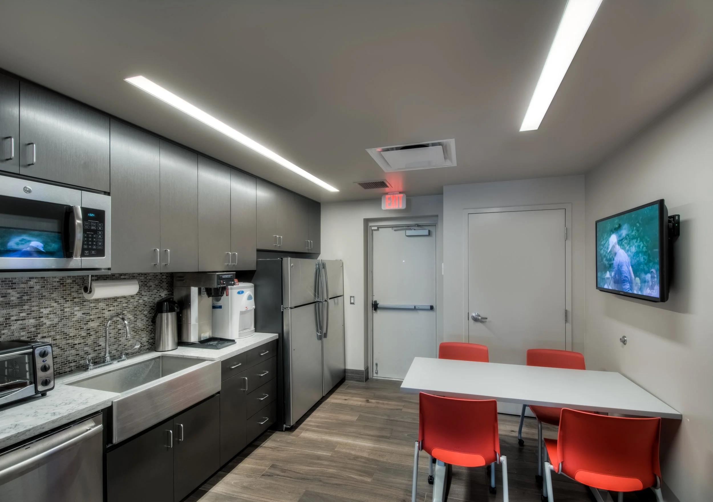 Modern kitchenette with stainless steel appliances, a microwave, coffee makers, a sink, and a small table with four red chairs in a well-lit room.