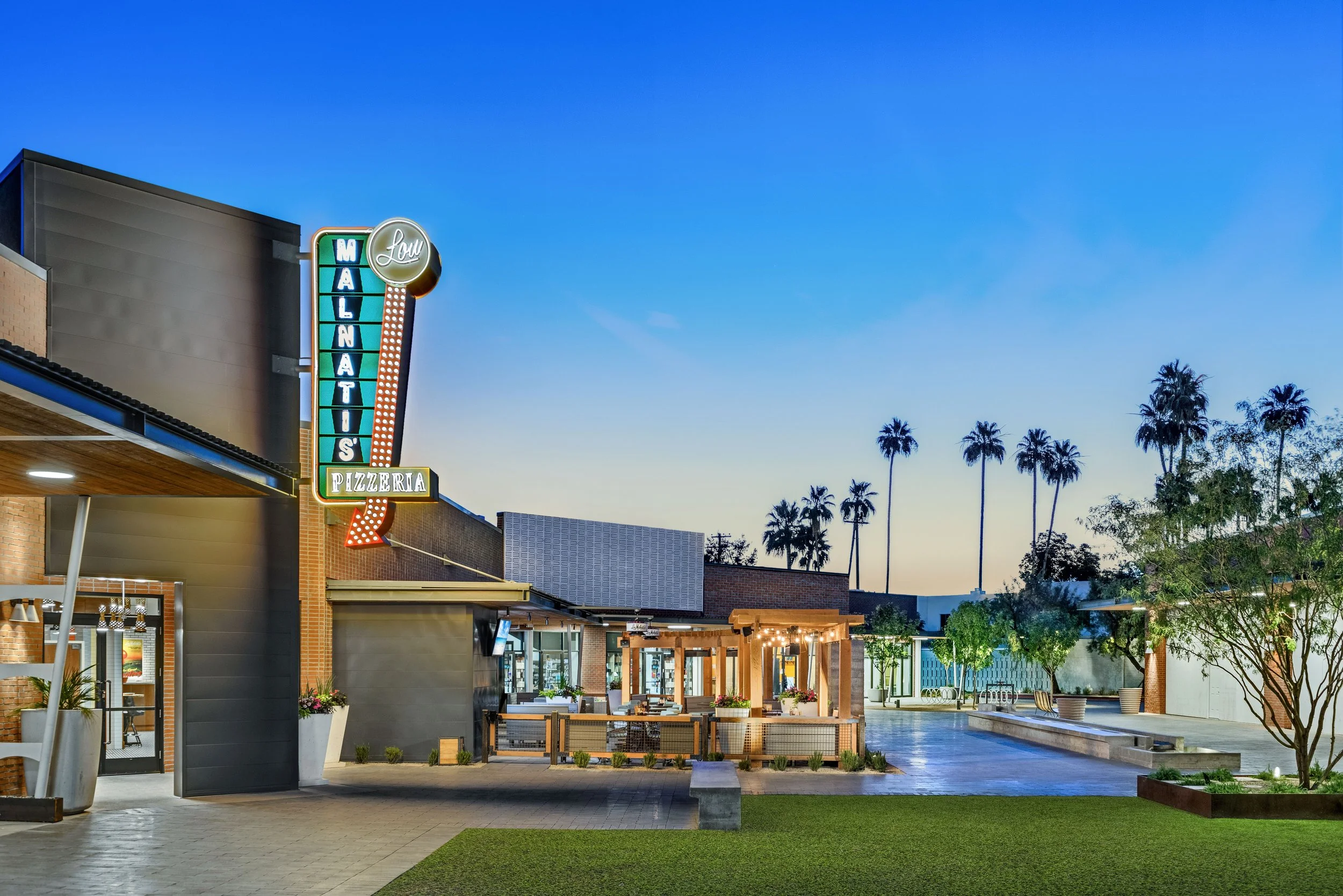 Exterior view of a shopping and dining area with a neon sign for Malnat's Pizzeria, outdoor seating with string lights, trees, and palm trees against a clear evening sky.