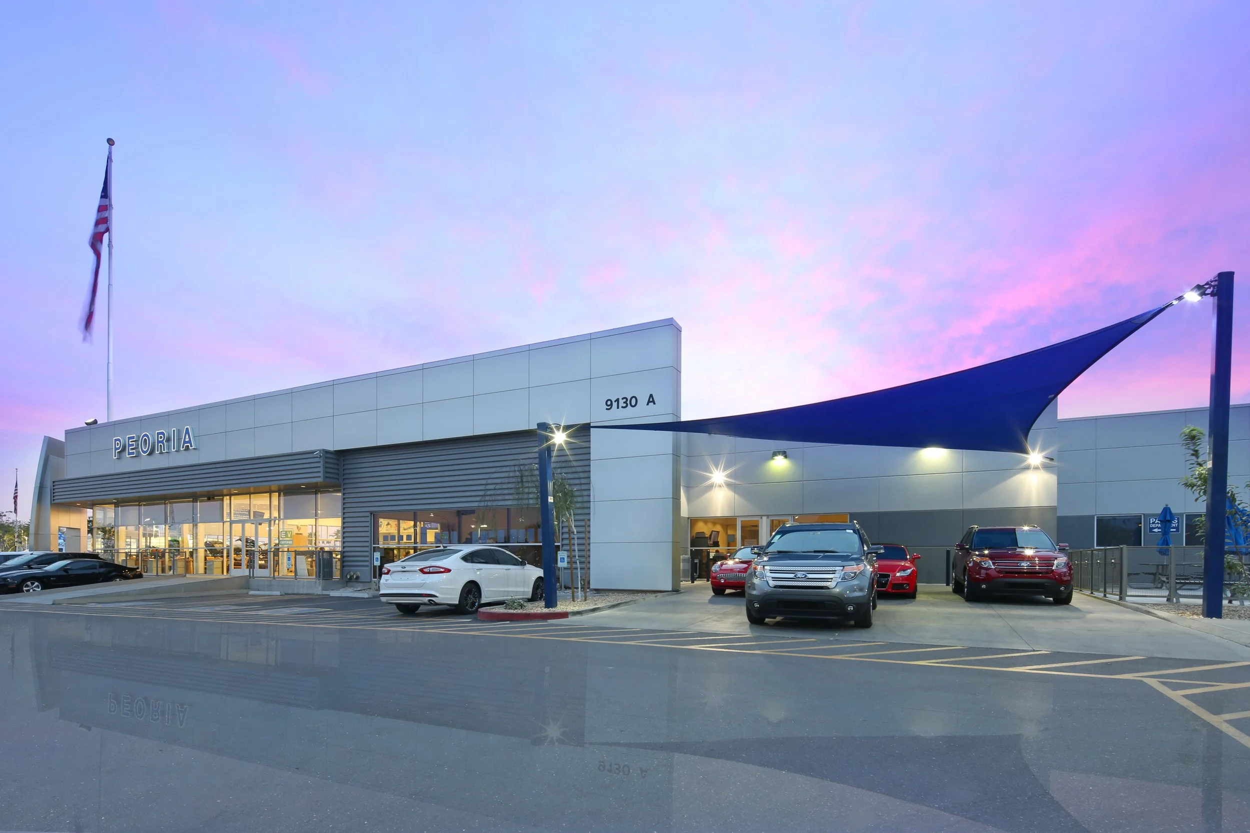 Front view of Peoria dealership building with cars parked outside, blue shade sail overhead, American flag, and a sunset sky in the background.