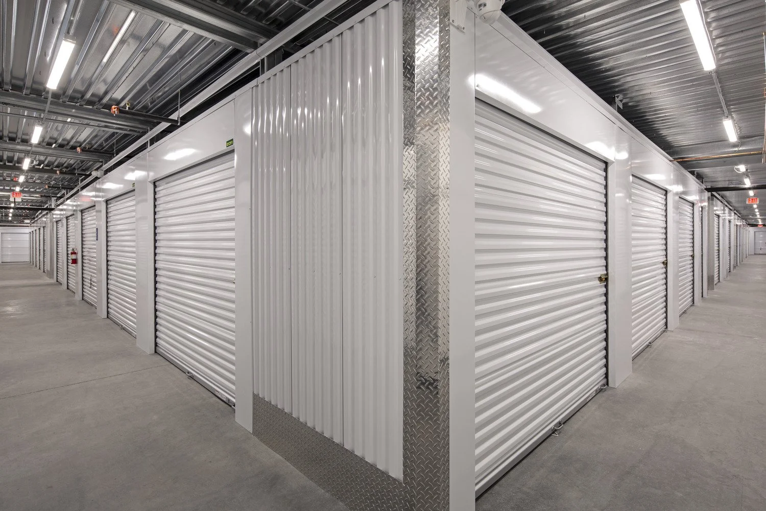 Interior view of a storage facility with multiple rows of white roll-up storage unit doors and concrete floors.