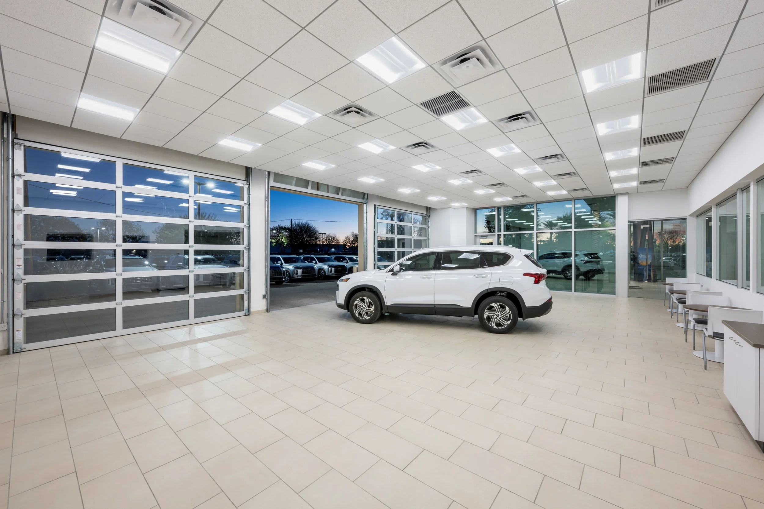 A white SUV inside a car dealership showroom with large glass windows and garage doors, several other cars visible outside, modern ceiling lights, tiled floor, and chairs along the wall.
