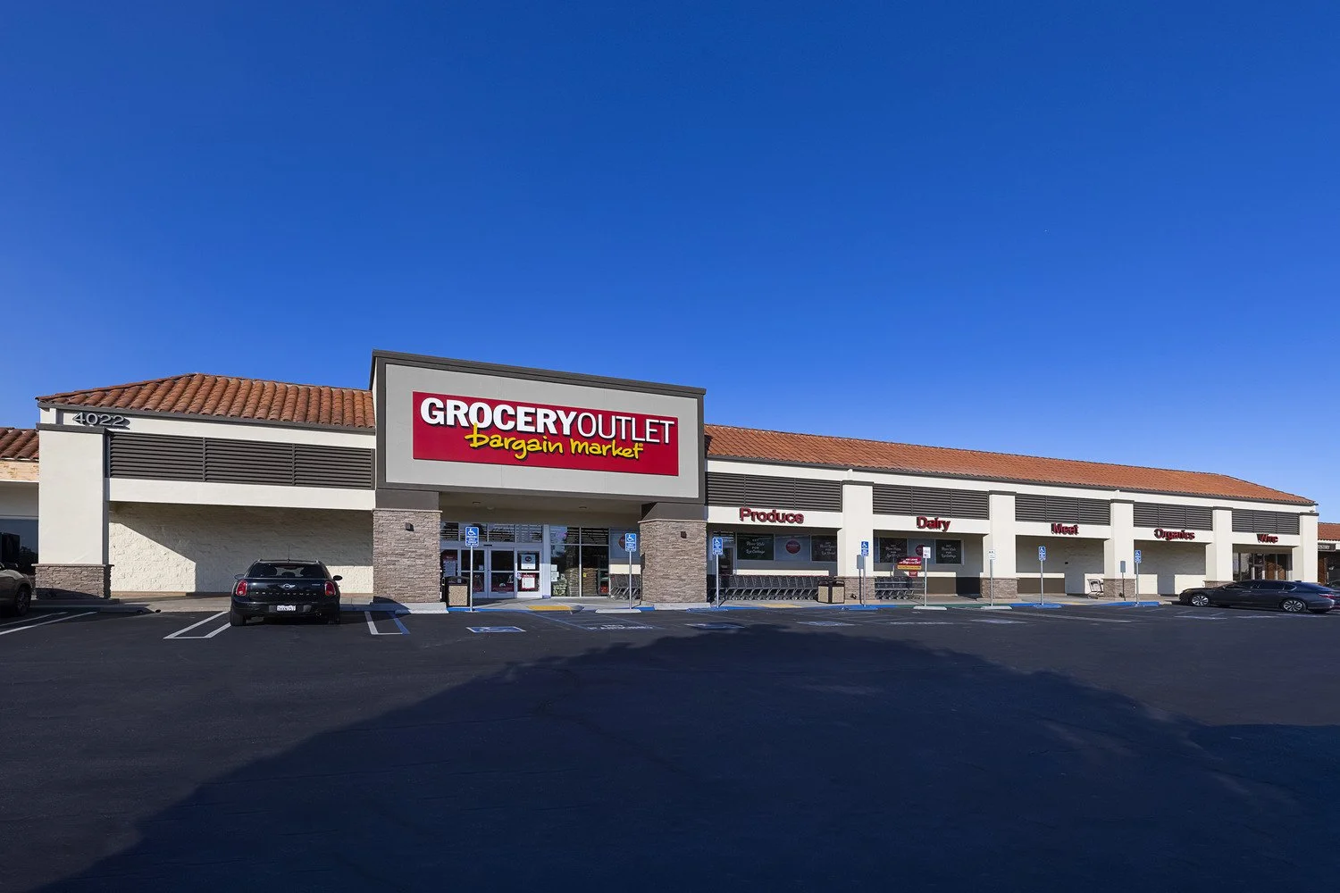 Exterior view of a grocery store named Grocery Outlet Bargain Market with a large red sign, a beige building with a red-tiled roof, and a parking lot with cars under a clear blue sky.