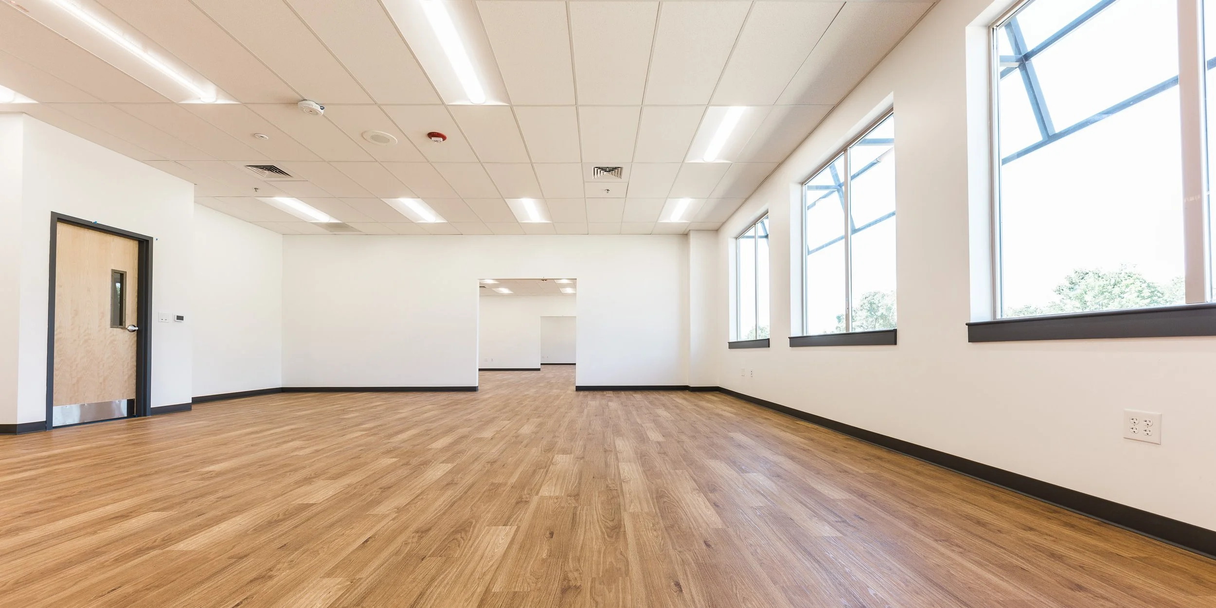 Empty office or conference room with wood flooring, white walls, a doorway, and large windows with a view of trees outside.
