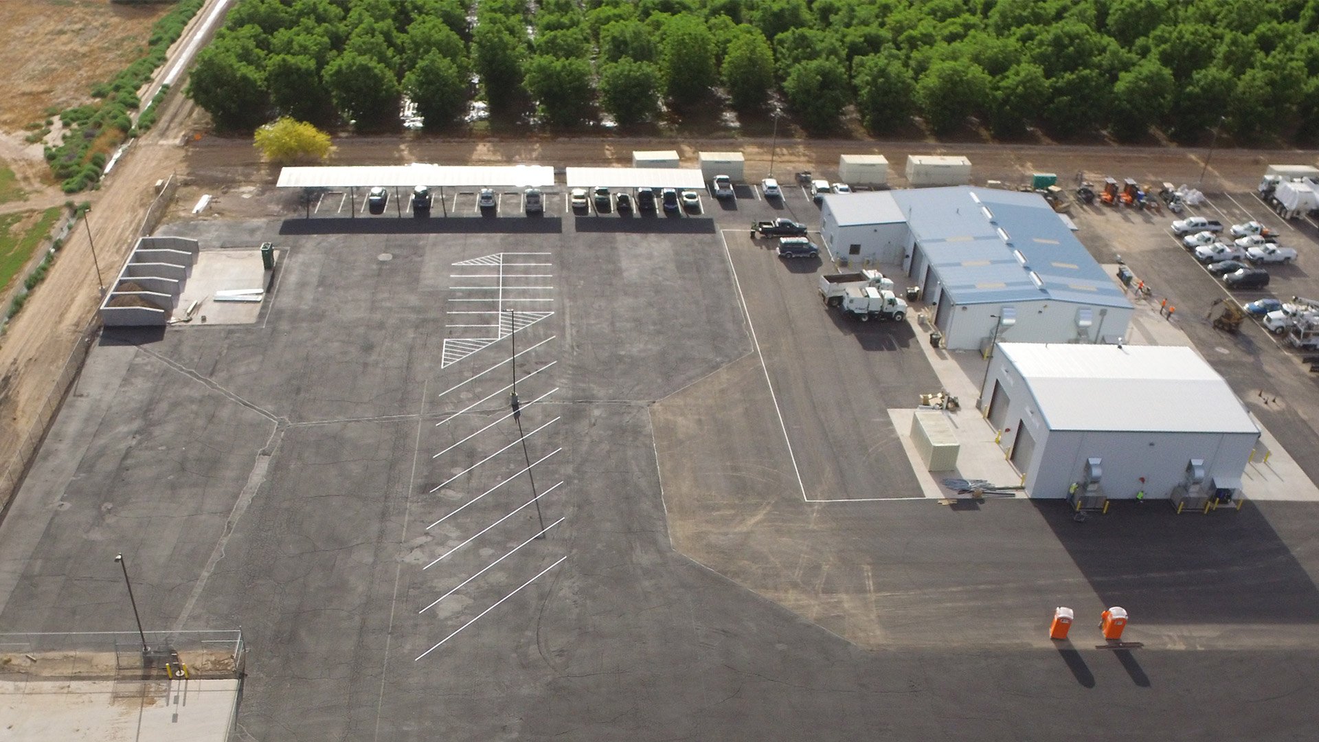A new parking lot with empty spaces and surrounding buildings, with some parked trucks and cars, and a background of green trees.