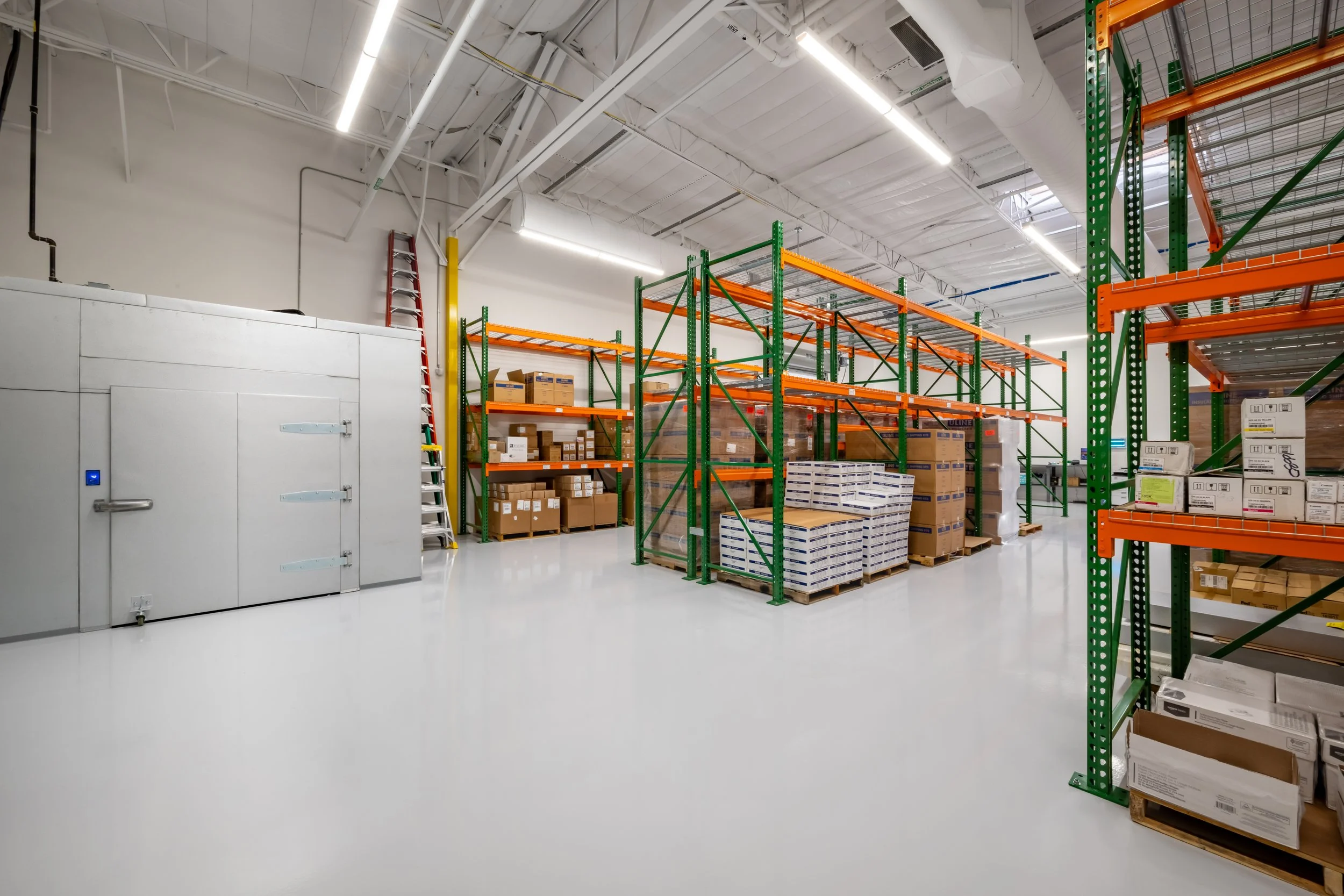 Empty warehouse with green and orange metal shelving units stocked with boxes, a ladder leaning against the wall, and a large cold storage door on the left.