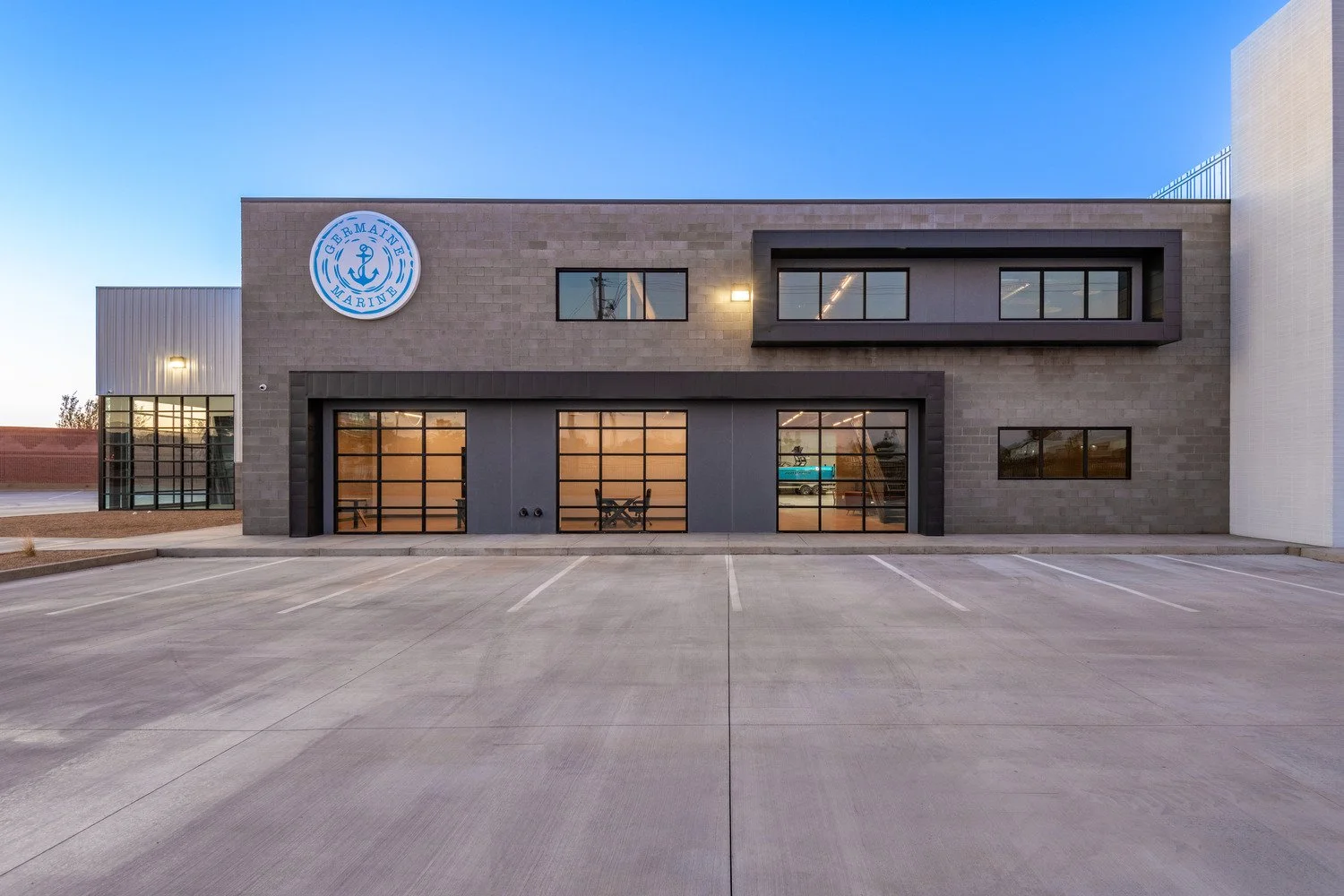 Modern commercial building with a sign that reads 'Germain Marine', glass garage doors, empty parking lot, and a clear blue sky.