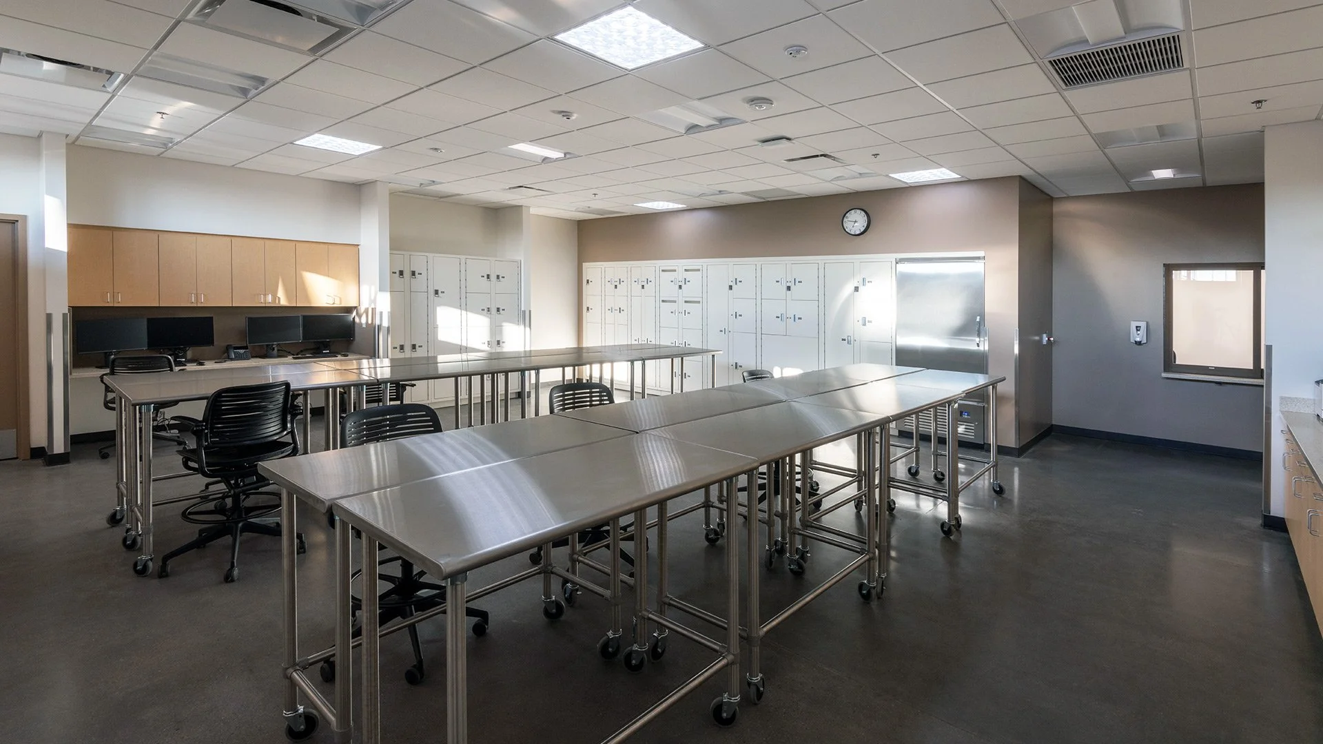 Empty classroom with stainless steel tables, black chairs, wall-mounted lockers, a clock, computer monitors, and a small window.