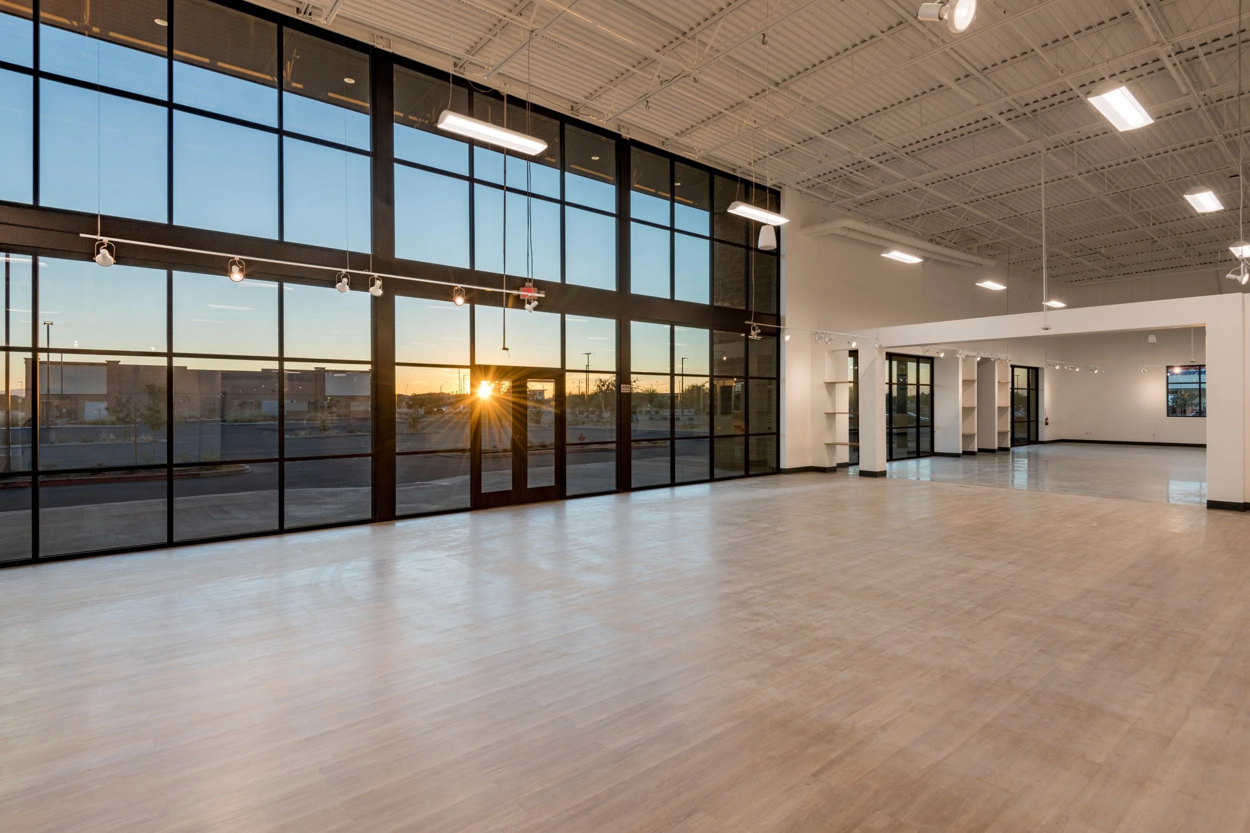 Empty commercial space with large glass windows showing sunset, polished wood floors, white walls, and high ceiling with industrial lighting.