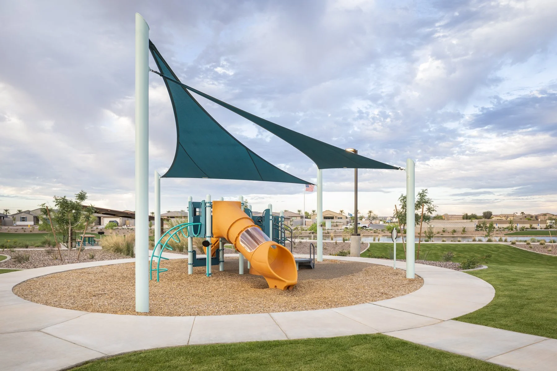 Empty playground with a blue shade sail, orange slide, and concrete walking paths in a neighborhood park with houses and trees in the background.
