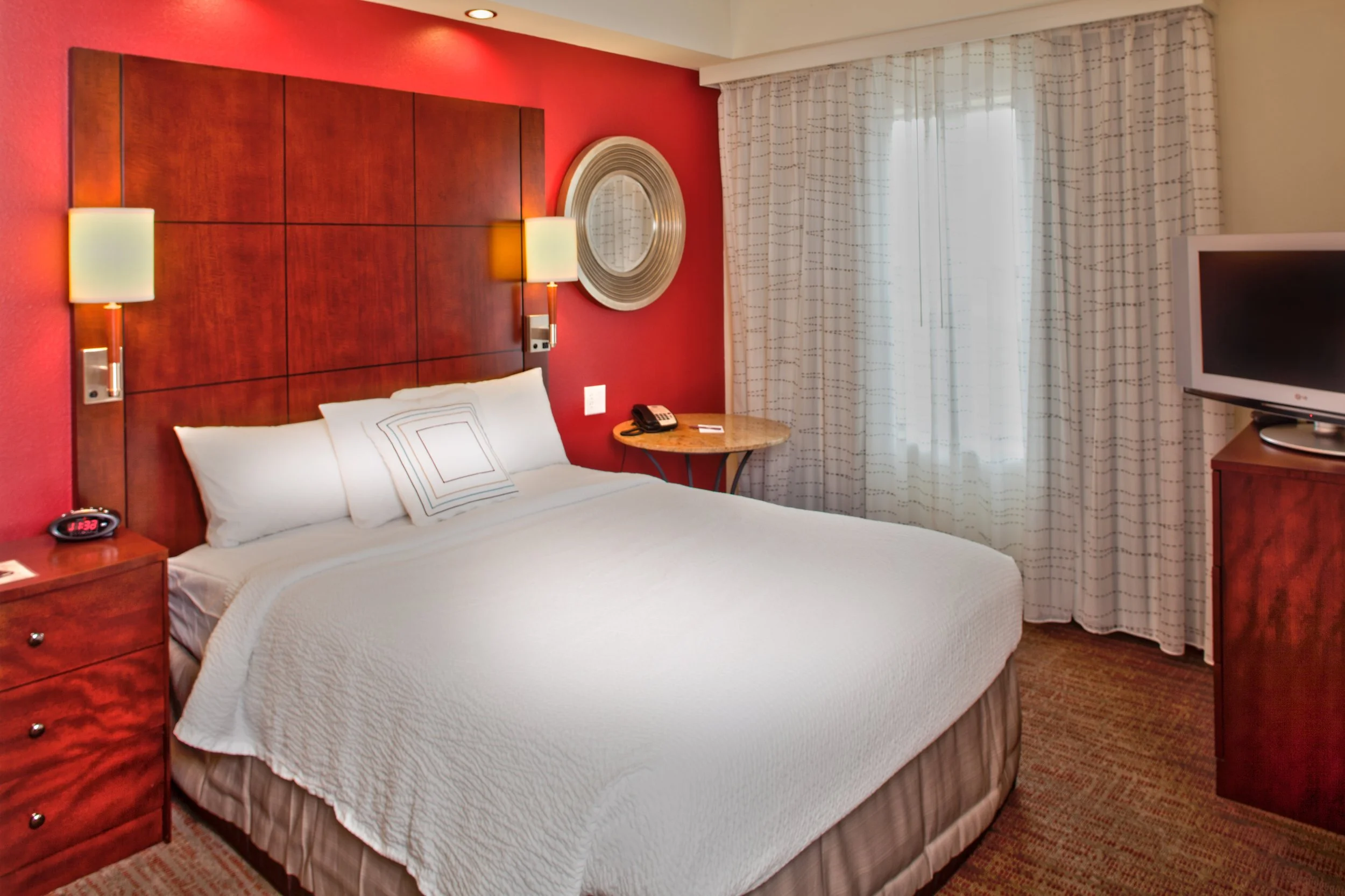 A hotel room with a large bed, white bedding, and pillows, against a red accent wall with a wooden headboard. There are two wall-mounted lamps on either side of the bed, a round mirror on the wall, a window with white curtains, a bedside table with a