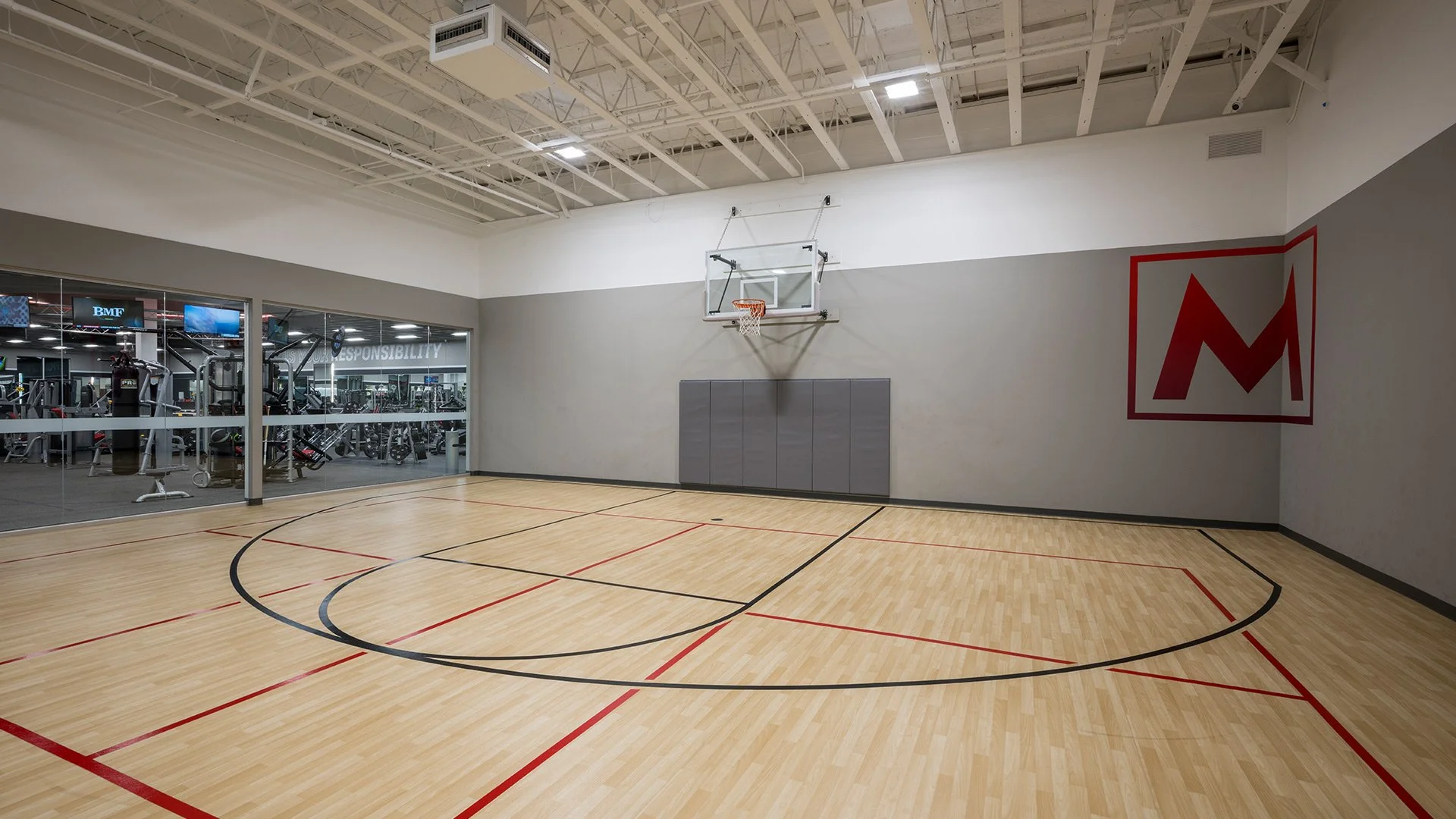 Empty indoor basketball court with wooden flooring, basketball hoop, and a gray padded wall, adjacent to a gym visible through glass. There is a large red and black logo on the right wall.