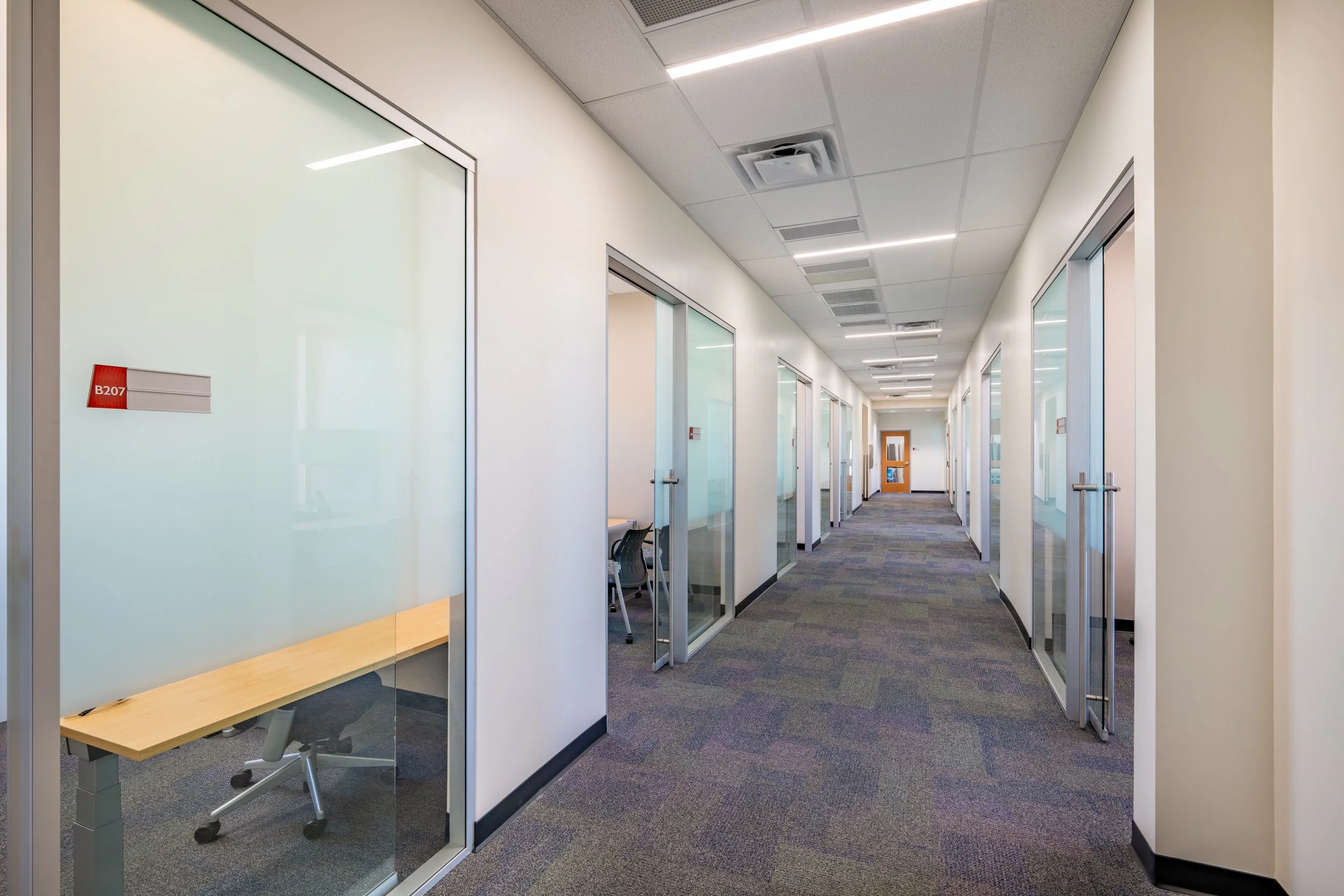 Empty office corridor with glass-walled offices on both sides and a wooden door at the end.