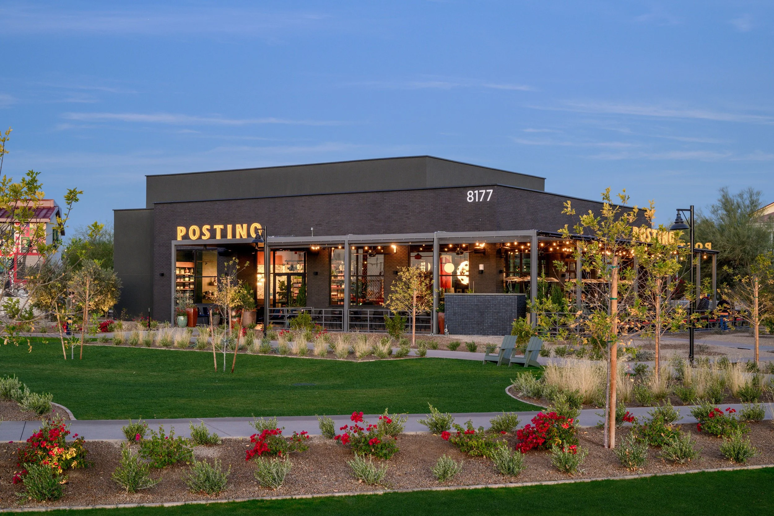 A modern black-brick restaurant building with illuminated yellow signs reading 'POSTING' at night, surrounded by landscaped garden with trees, shrubs, and flowers.