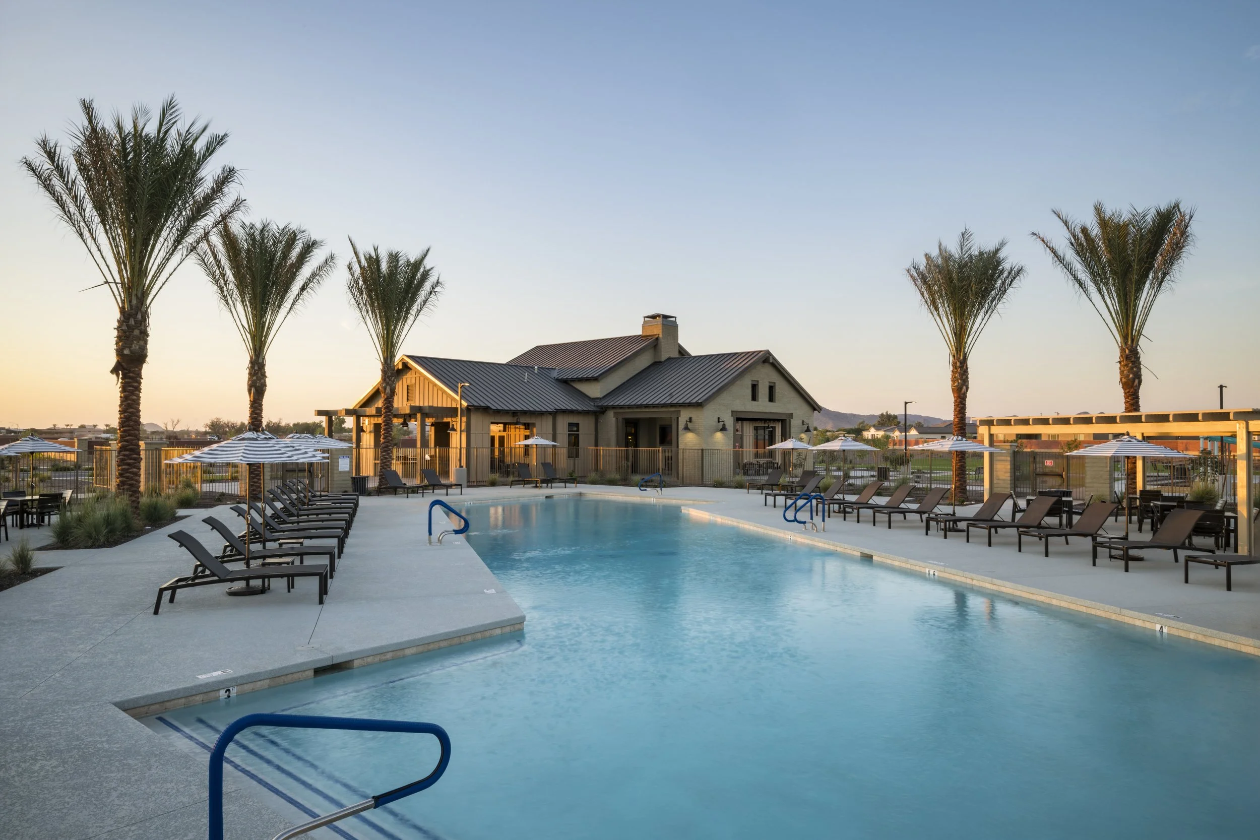 Outdoor swimming pool with lounge chairs, umbrellas, and palm trees at sunset, near a house or clubhouse.
