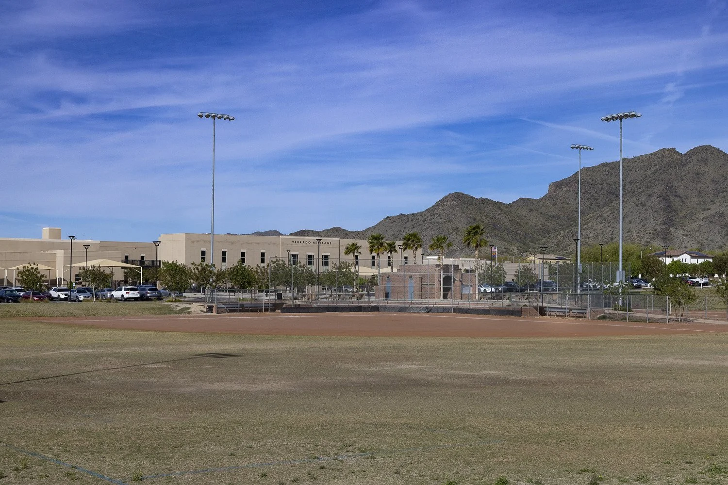 A sports field with a dirt infield, grass outfield, and flagpoles under a blue sky, with mountains and a school building in the background.