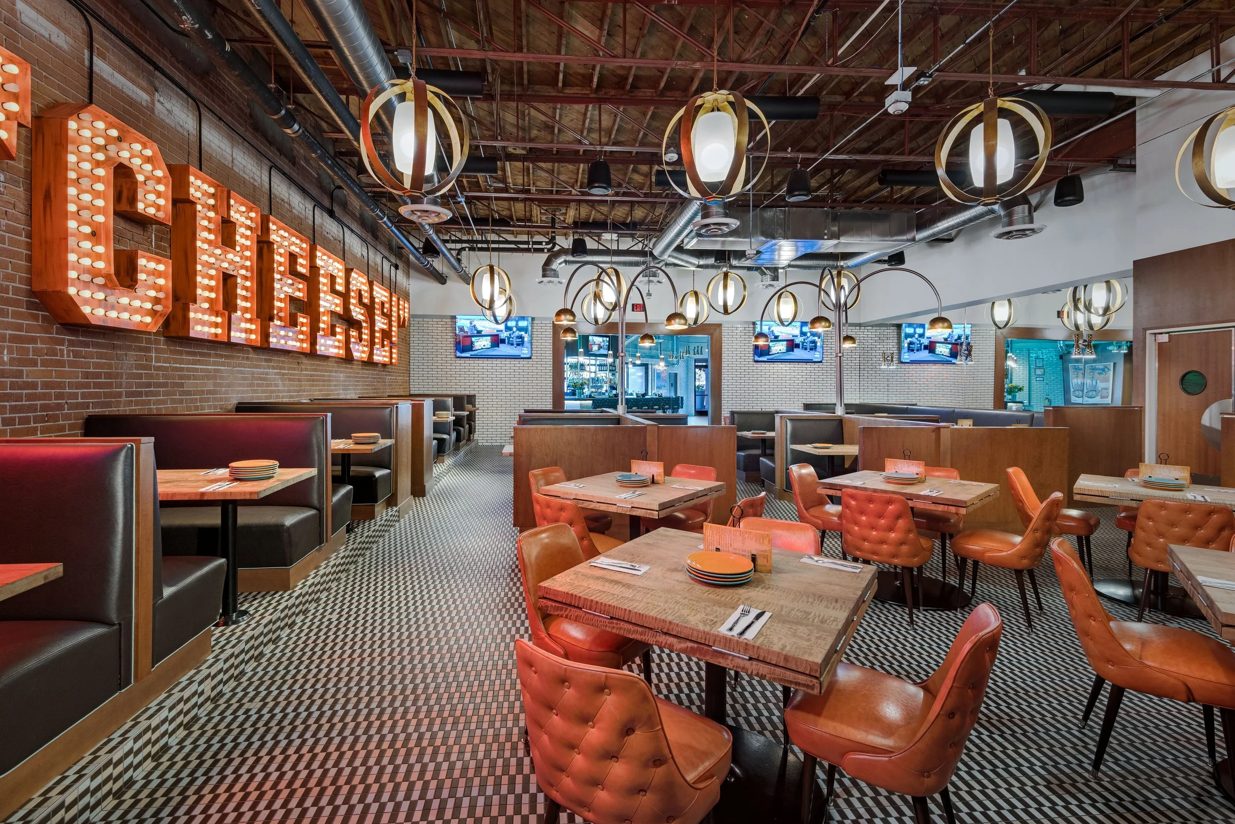Empty restaurant with wooden tables and leather chairs, brick walls, decorative hanging lights, and neon sign that reads 'CHEESE' on the wall.
