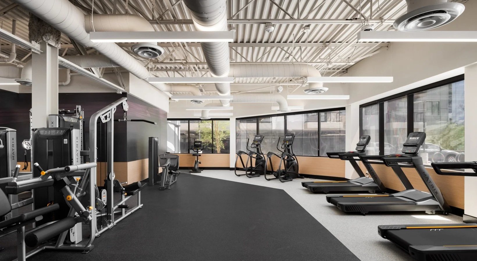 Empty modern gym with cardio machines, treadmills, ellipticals, and exercise equipment near large windows.