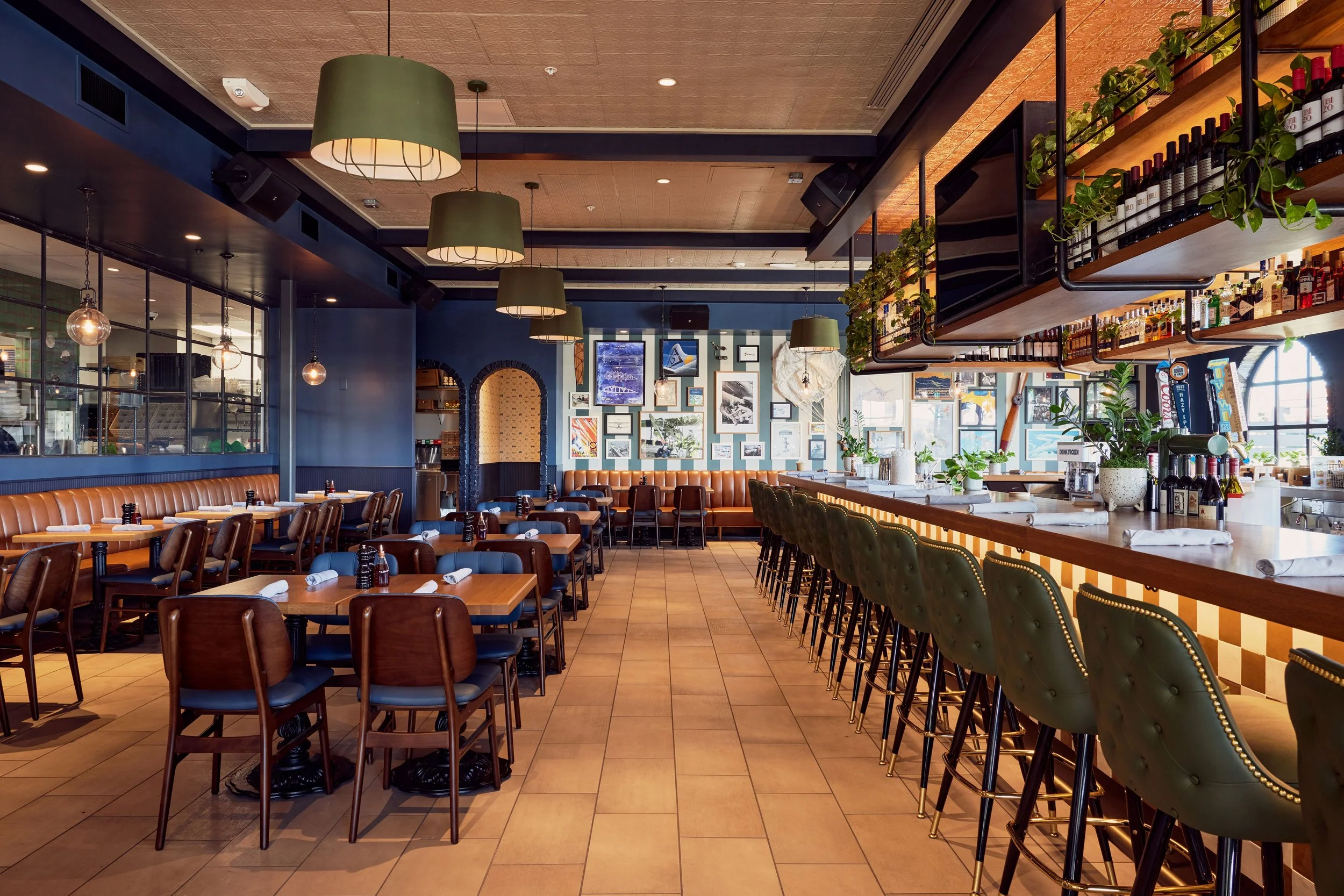 Inside of a modern restaurant with a bar counter, green barstools, and hanging pendant lights, decorated with framed art on the wall and potted plants.