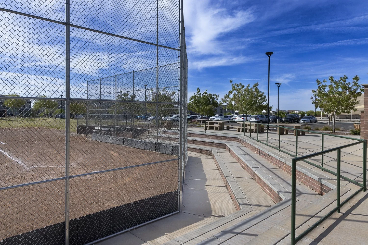 A baseball field enclosed by a tall chain-link fence, with dirt infield and grass outfield, adjacent to a paved walkway with brick steps, green rails, and a parking lot with cars and trees in the background under a blue sky with wispy clouds.