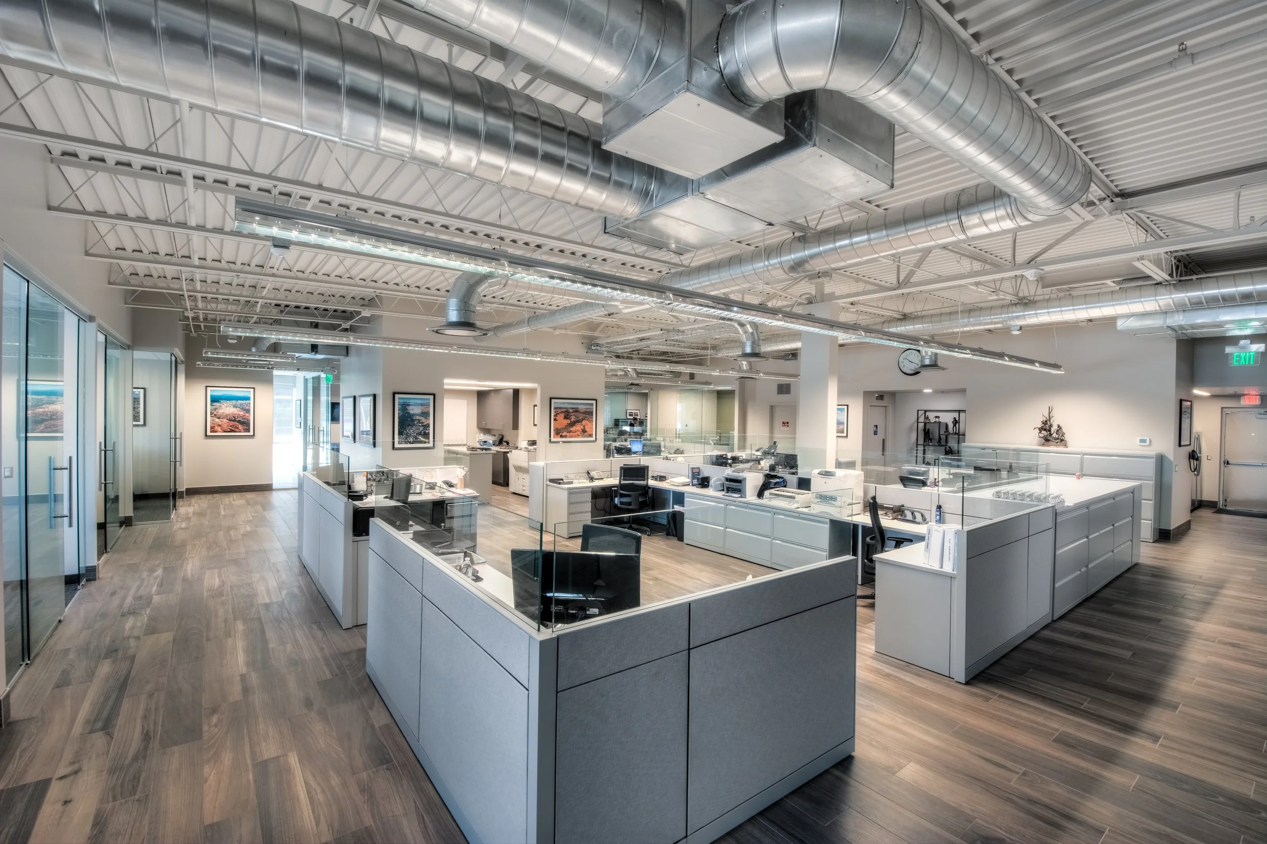 Modern open-plan office with workstations, glass partitions, ceiling ventilation ducts, and artwork on the walls.