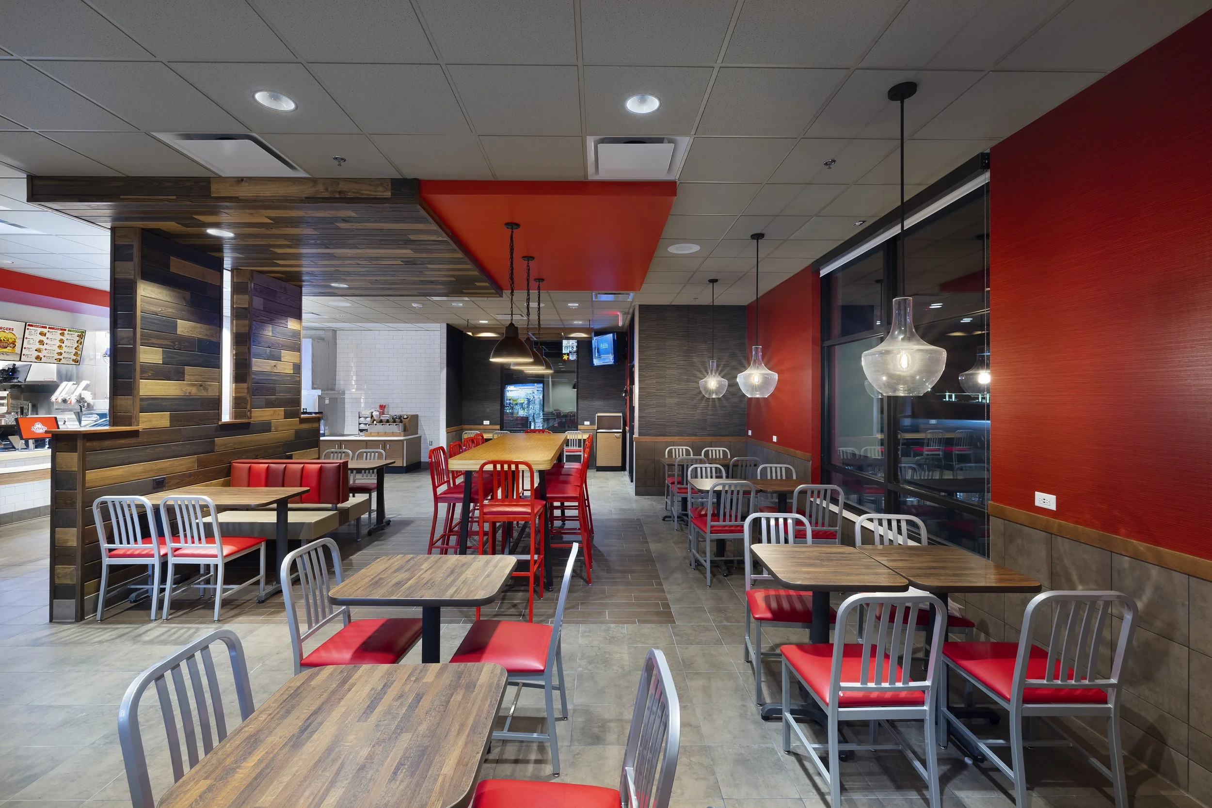 Interior of a fast-food restaurant with wooden and red accent decor, tables, and chairs, including pendant lights and a menu display at the counter.