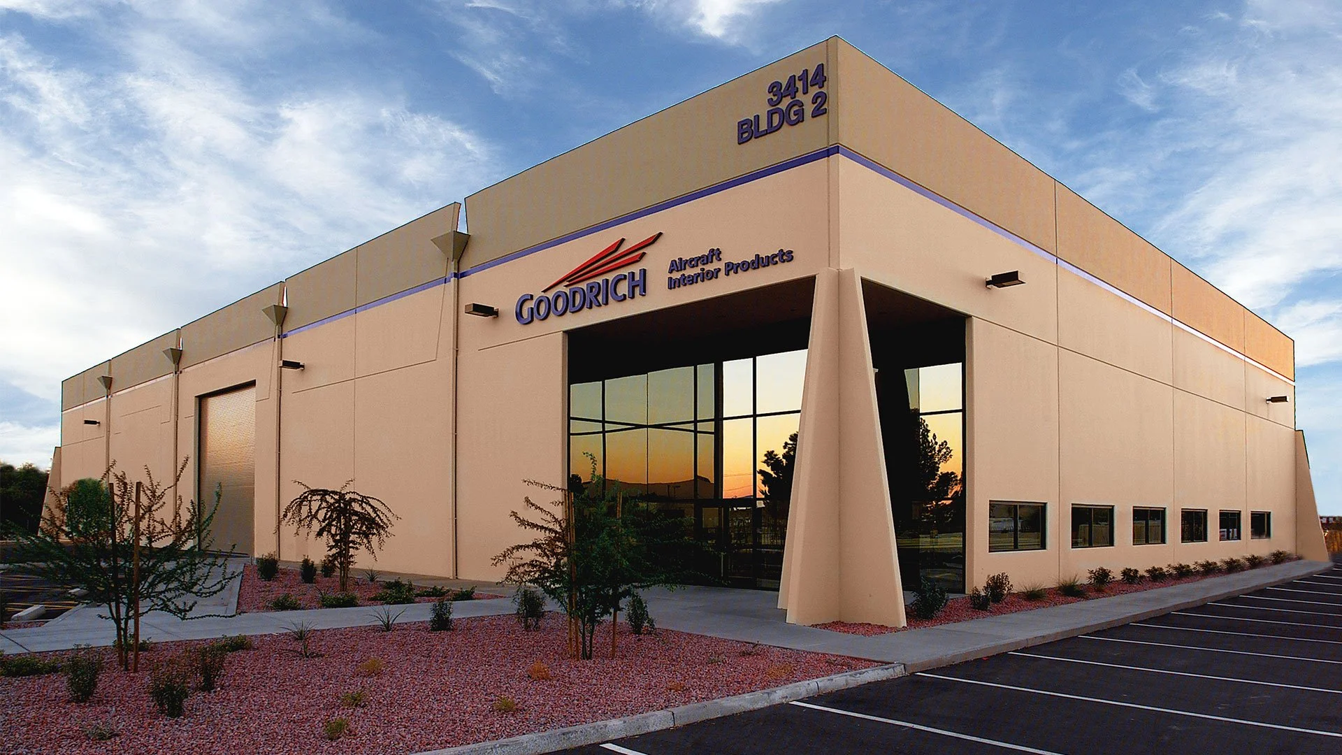 Exterior view of a large beige industrial building with blue signage reading 'Goodrich Aircraft Interior Products.' The building has a glass entrance, small windows, and is surrounded by a landscaped area with small bushes and trees. There are parkin