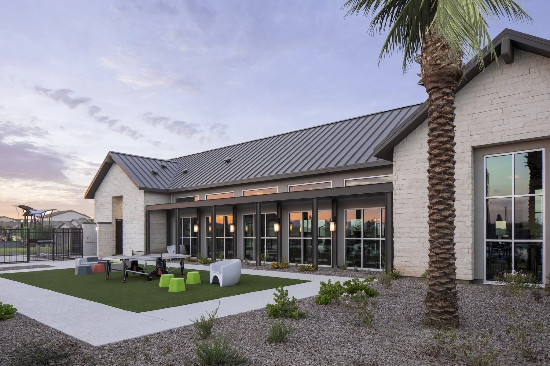 A modern house with a gray metal roof, large glass windows, a small backyard featuring a green artificial turf area with colorful outdoor furniture, and a tall palm tree. Sunset sky in the background.